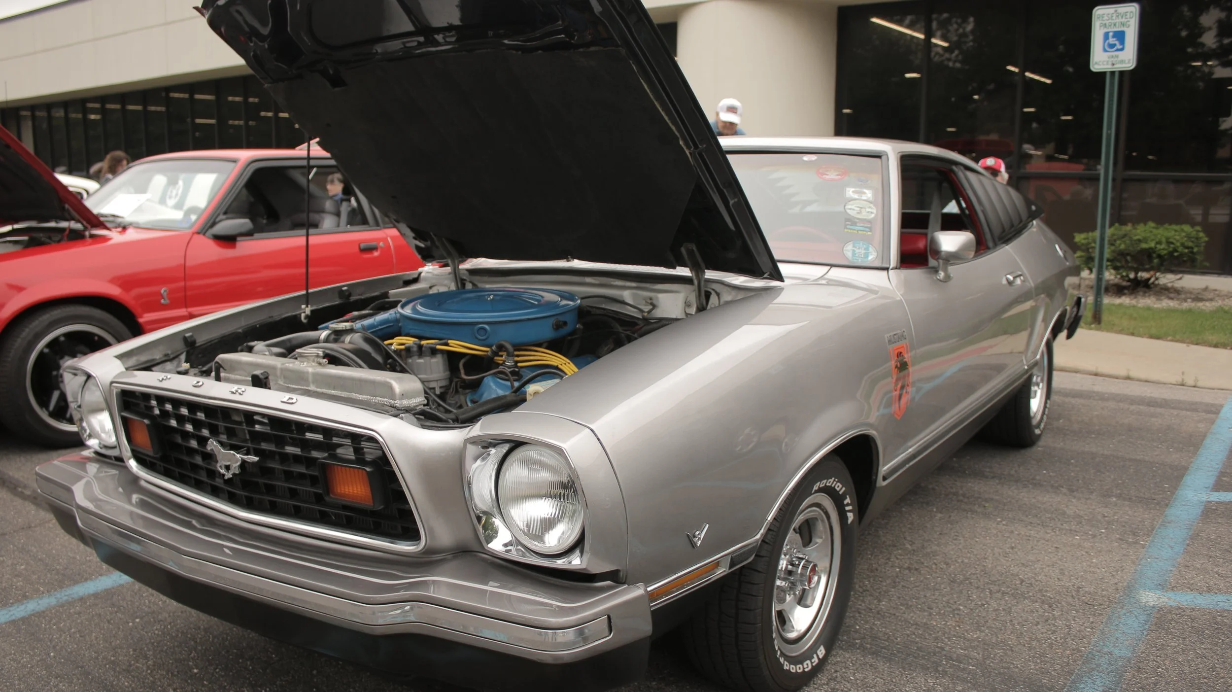 A silver classic Ford Mustang with its hood open on display at a car show. Next to it is a red car, and there are people in the background, some wearing caps, near a building with glass windows.