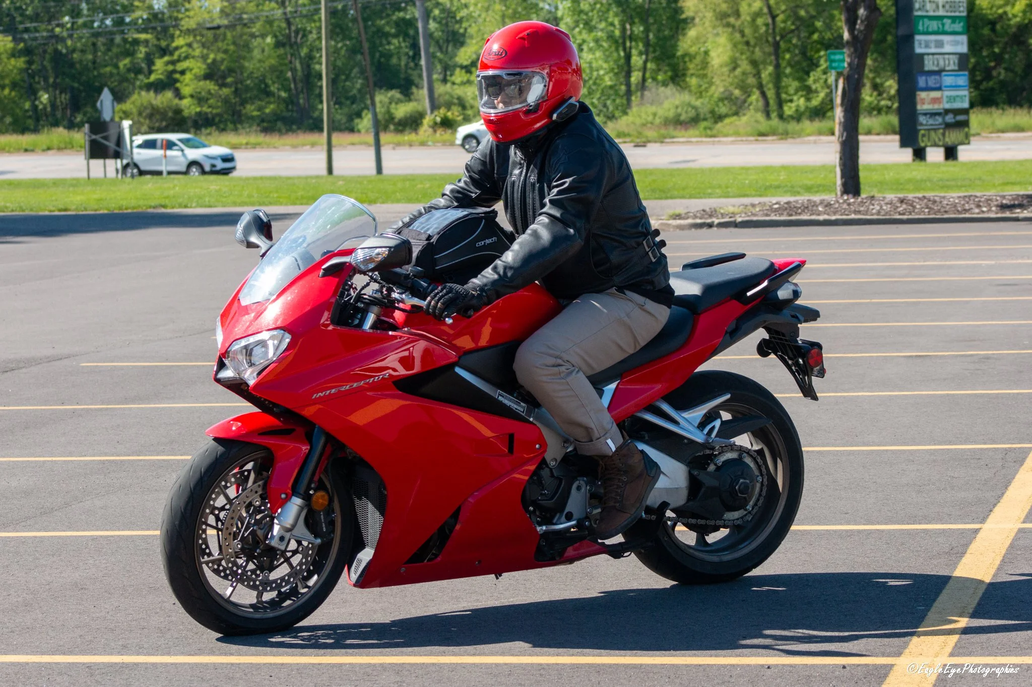 A person riding a red motorcycle in a parking lot, wearing a red helmet, black leather jacket, beige pants, and brown shoes.