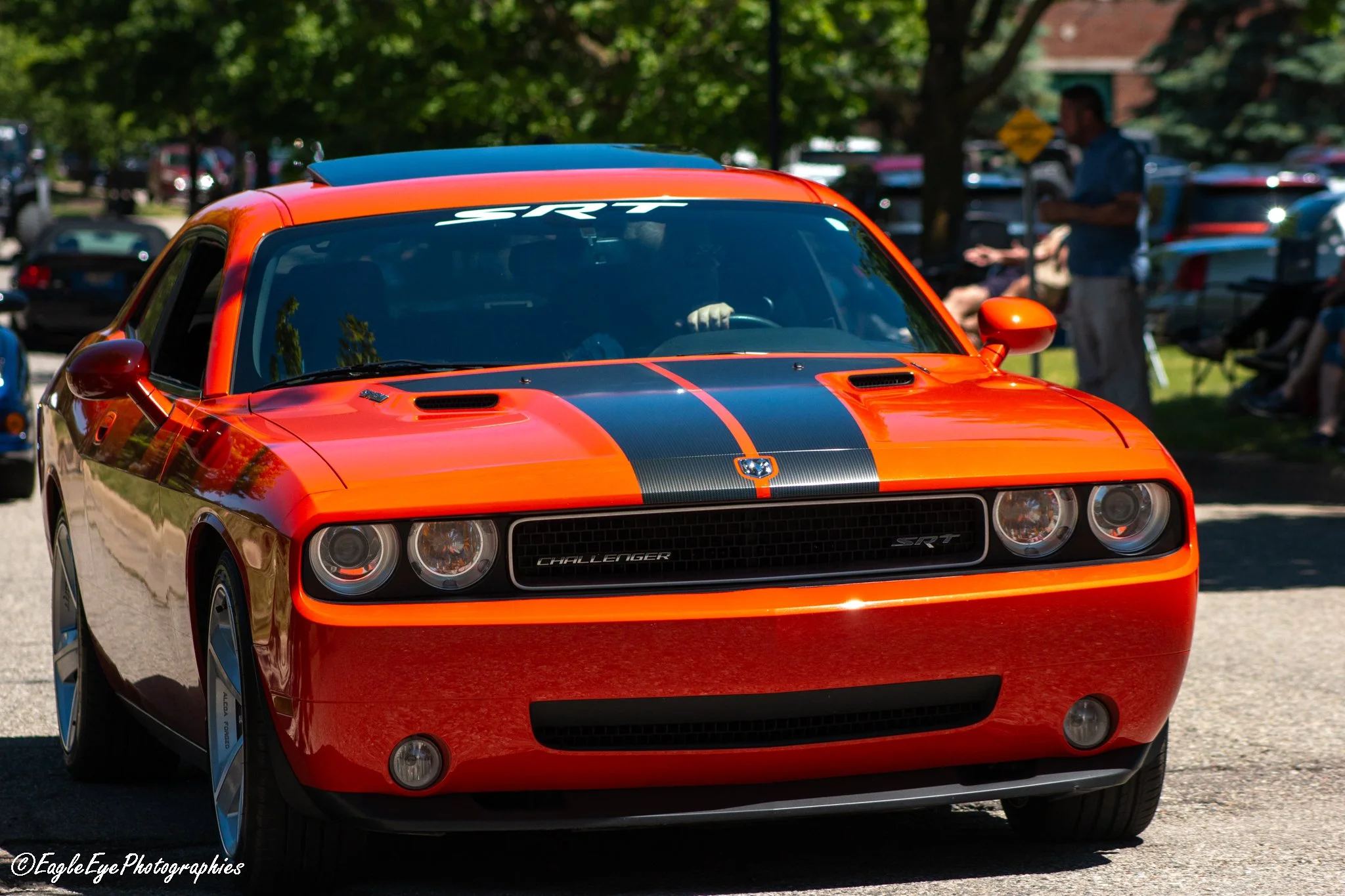 Close-up of a red Dodge Challenger SRT car with black racing stripes, parked outdoors at a car show with people and trees in the background.