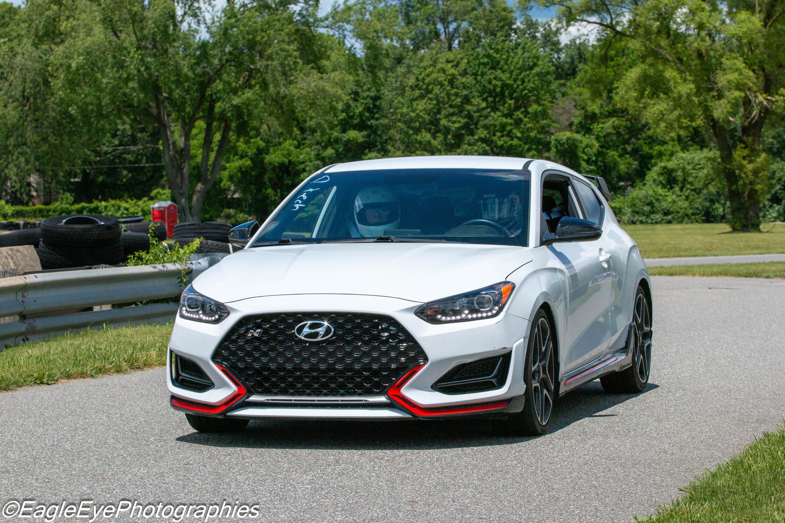 A white Hyundai Veloster N hatchback car parked on a street with trees and grass in the background, tire stacks on the side.