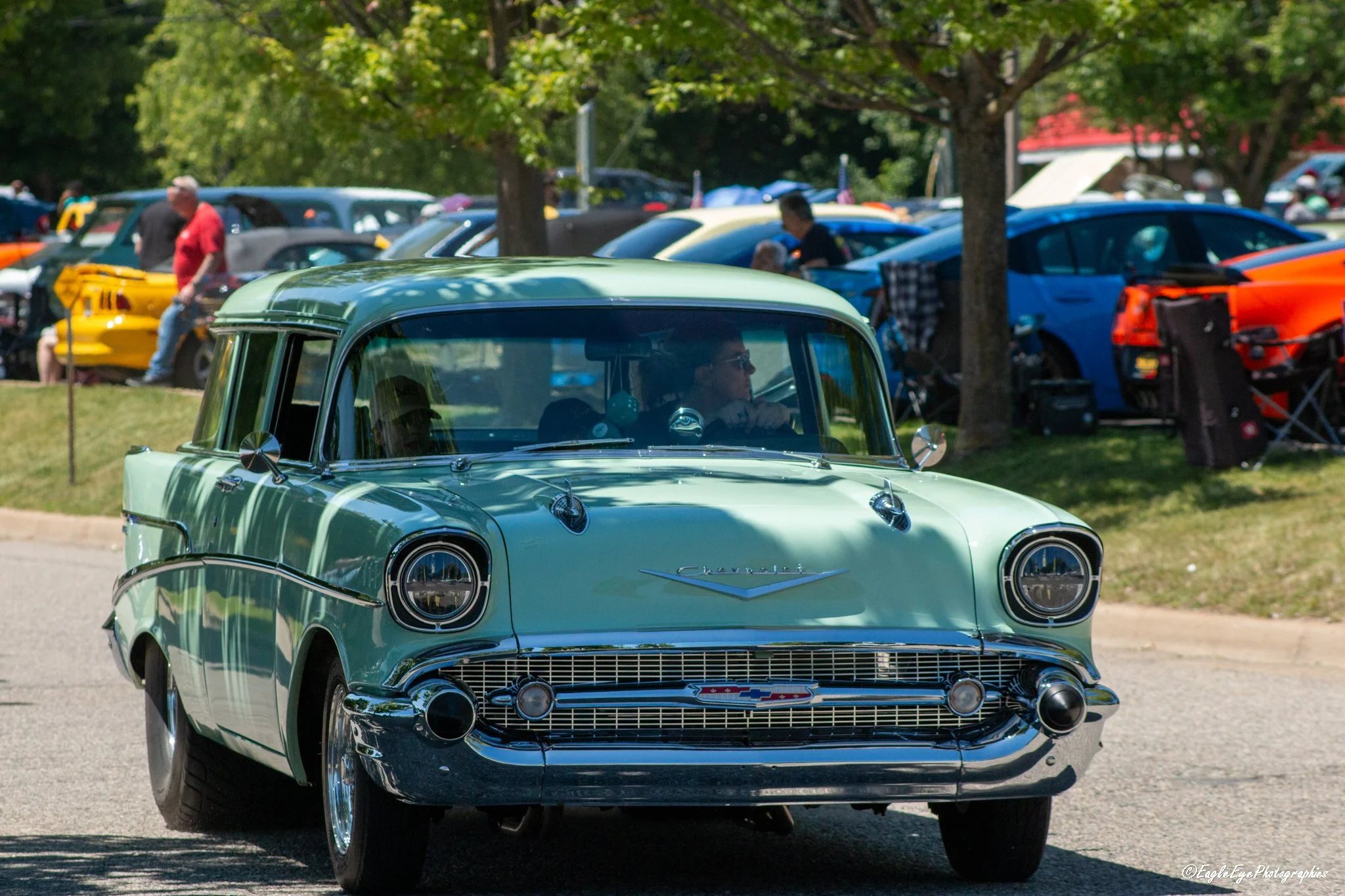 A mint green vintage Chevrolet car driving on a sunny day at a classic car show, with rows of colorful cars and people in the background.