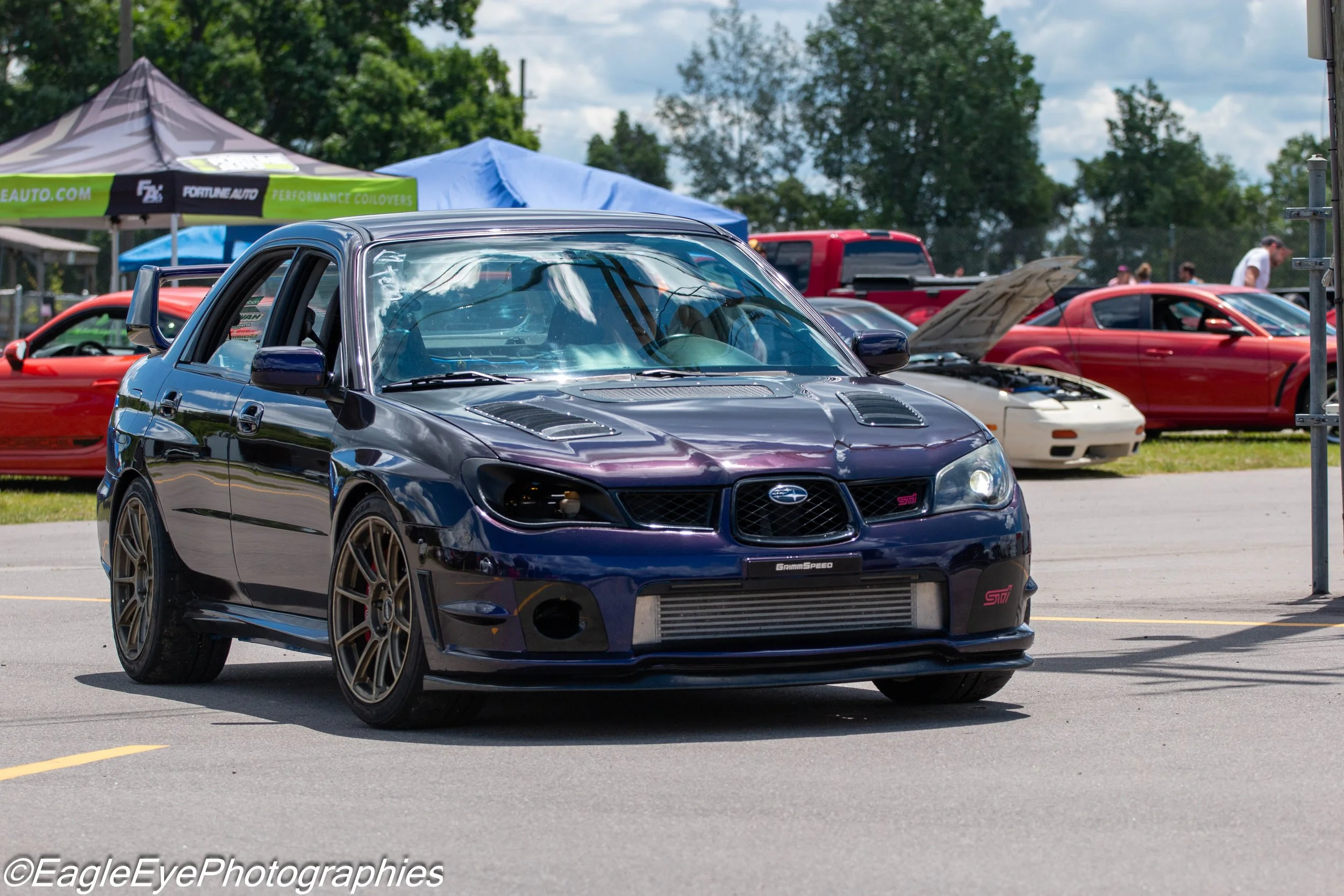 A black Subaru WRX STI with bronze wheels parked at an outdoor car show, with other cars and tents in the background.