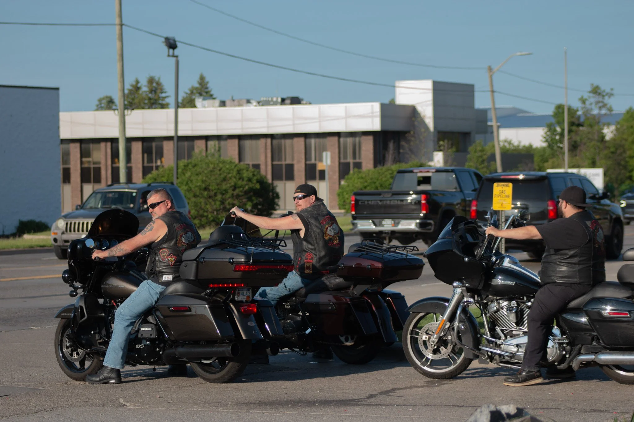 Bikers riding motorcycles on a street with buildings, trees, and utility poles in the background on a sunny day.
