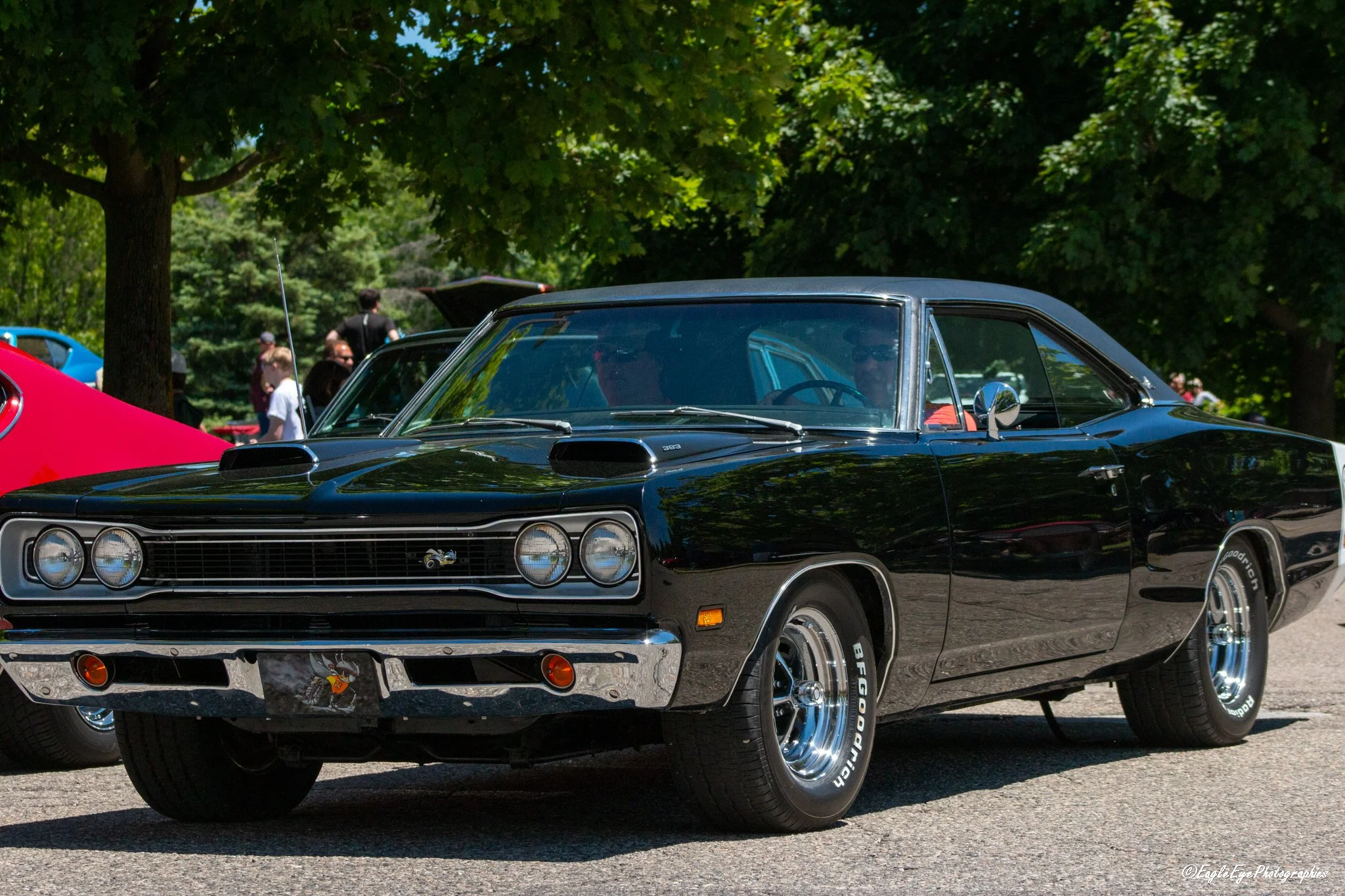 Black vintage muscle car parked outdoors with other vehicles and people in the background, surrounded by green trees.