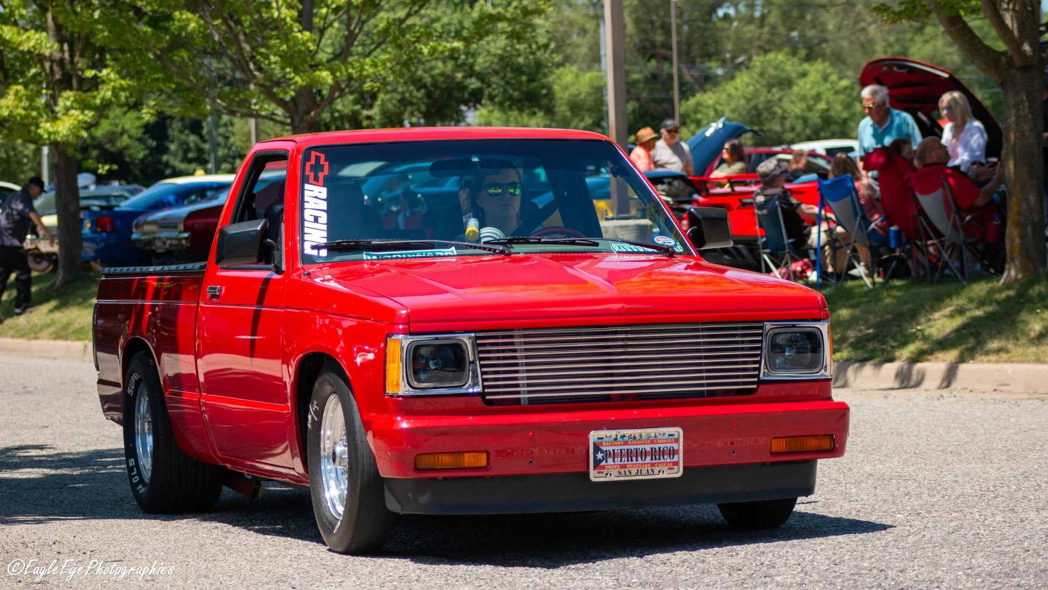 A red vintage pickup truck driving down a street during a daytime outdoor event surrounded by people and trees.