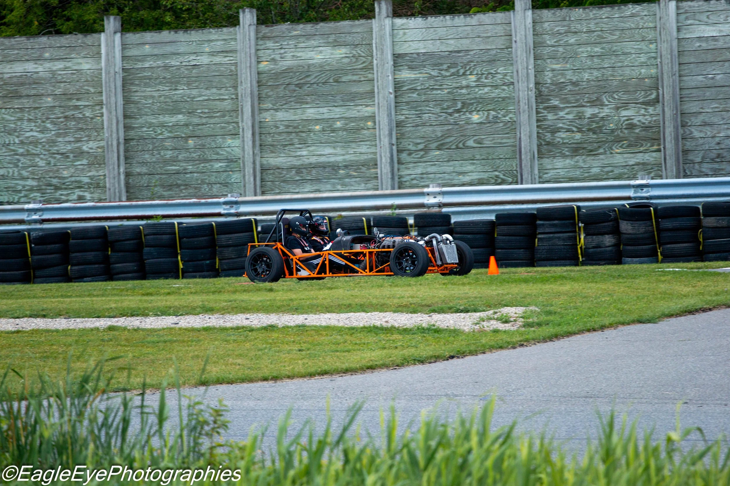 A small, open-wheel race car with two passengers wearing helmets, racing on a track bordered by tire barriers and a wooden fence.