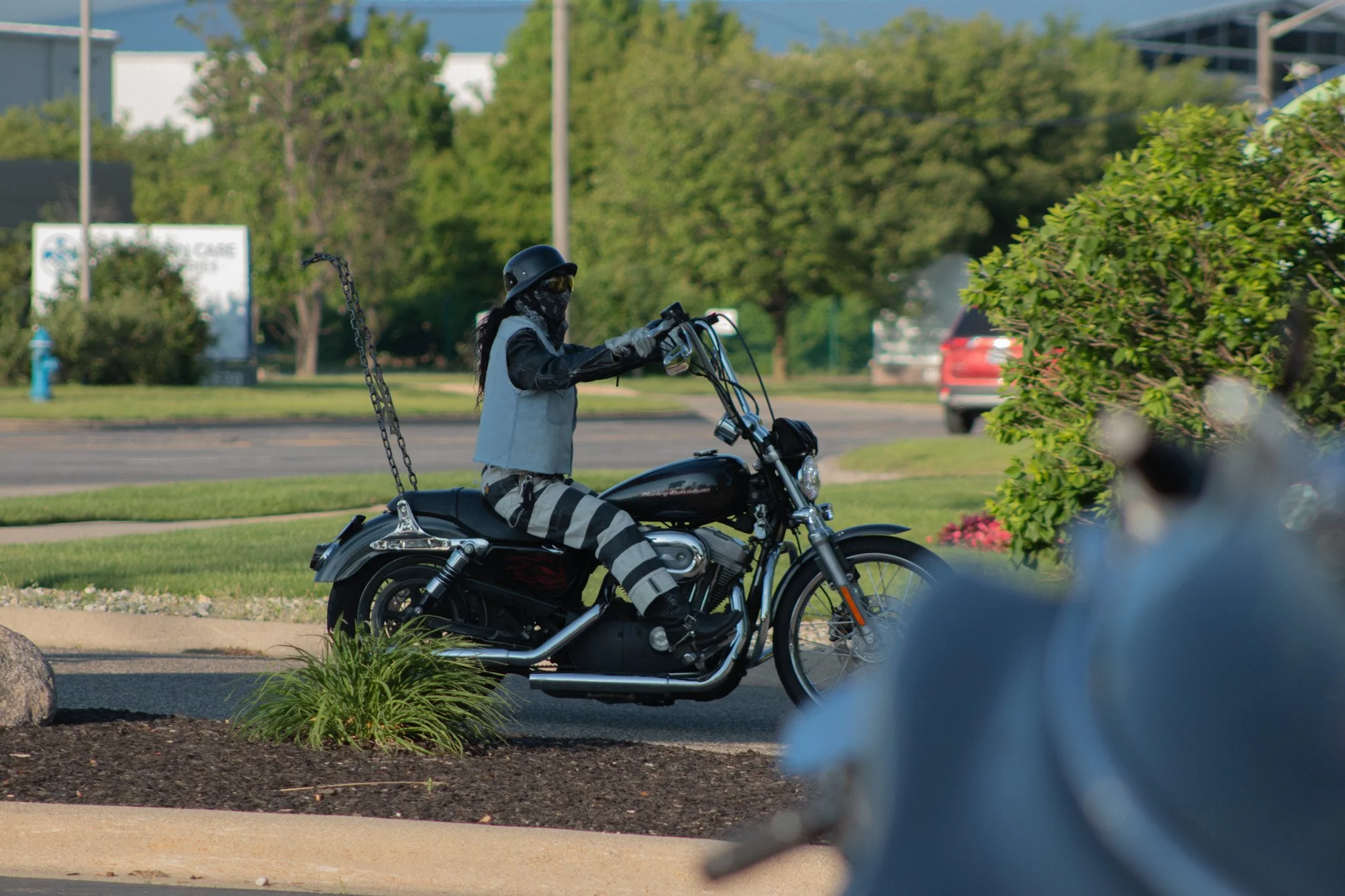 A person wearing a helmet, sunglasses, and a face covering riding a black motorcycle on a street with trees and a building in the background.