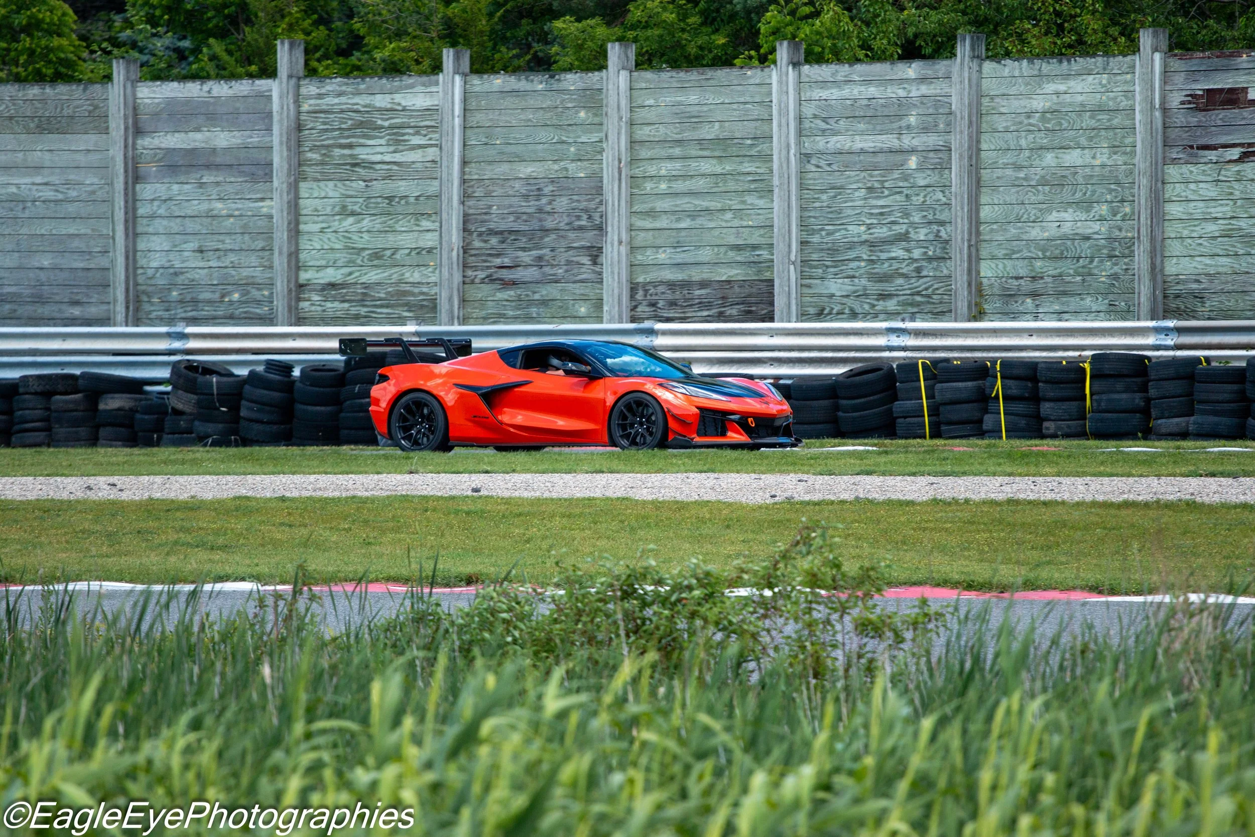 A bright orange race car parked on grass next to a stack of black tires with yellow straps, in front of a tall wooden fence and surrounded by green trees.