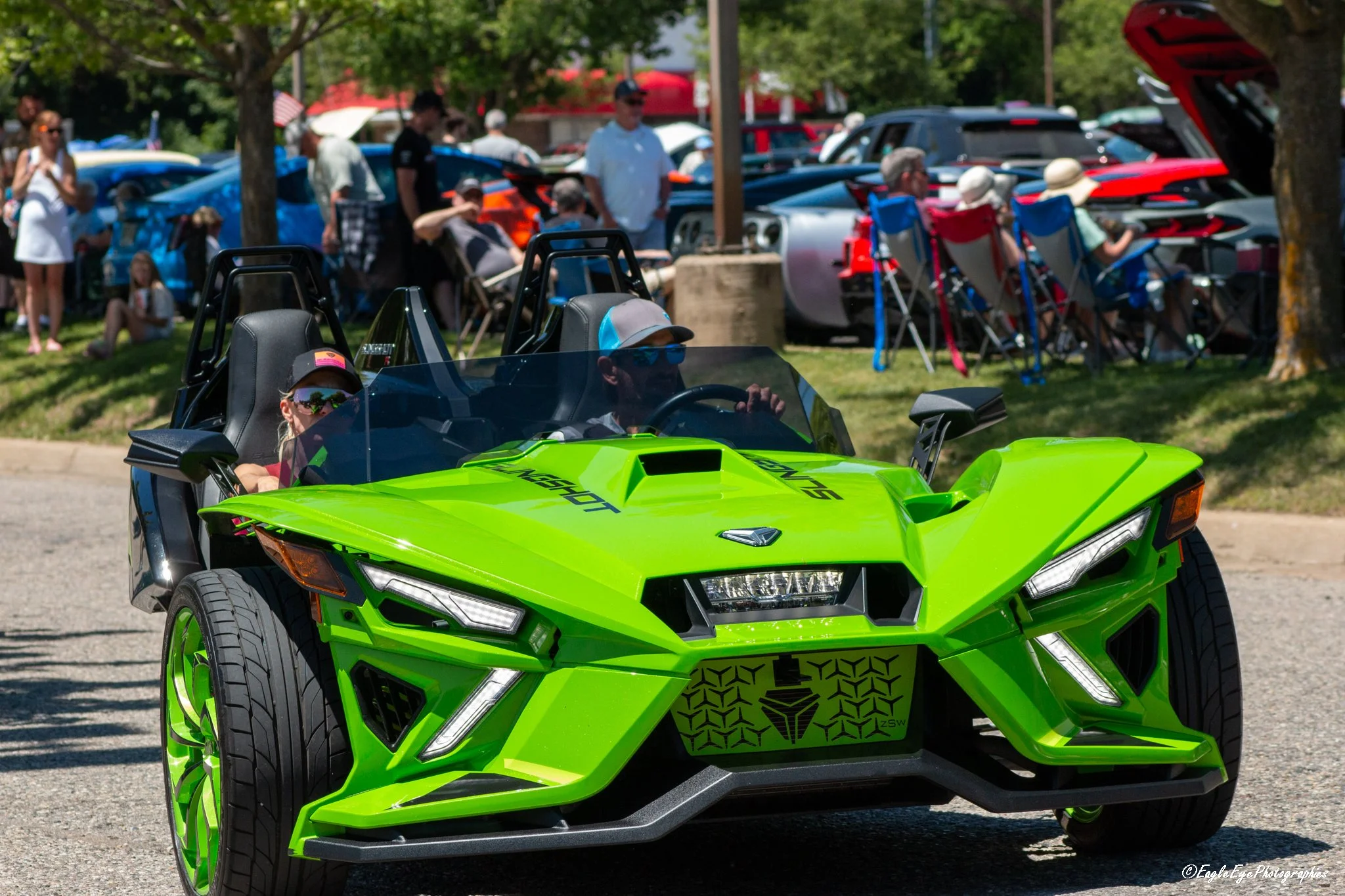 Green futuristic toy car with two children wearing hats and sunglasses seated inside, with a crowd of people and cars in the background at an outdoor event.