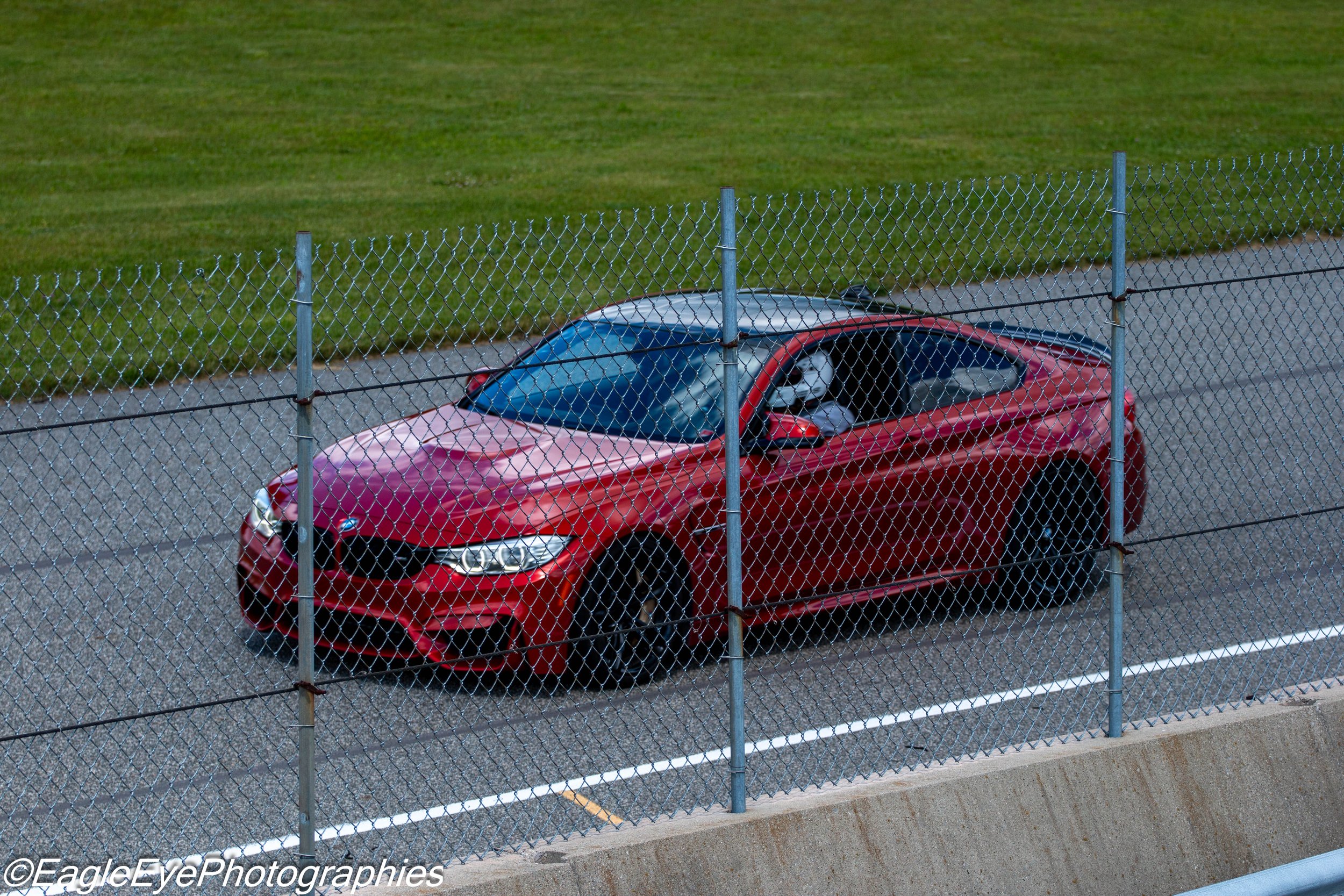 A red sports car parked behind a chain-link fence next to a grassy field.