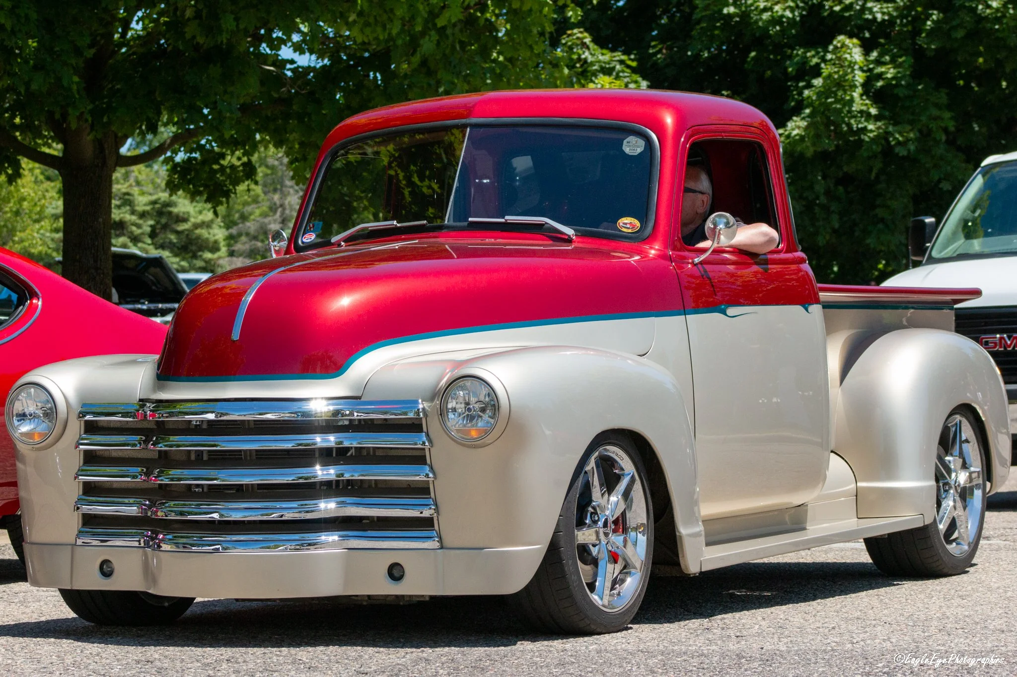 A vintage GMC pickup truck with a red and white paint job parked outdoors on a sunny day. There are trees in the background and a person with glasses in the driver’s seat.