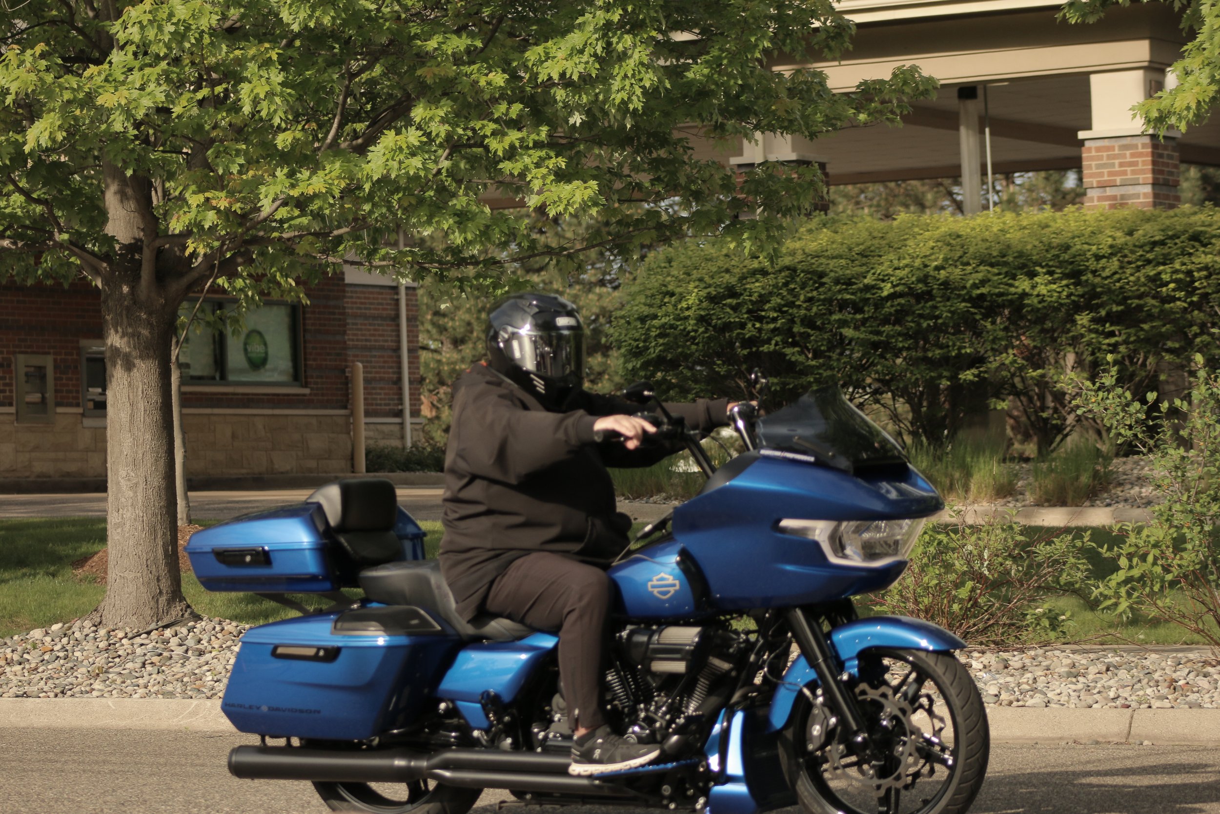 A person riding a blue Harley-Davidson motorcycle on a city street, wearing a black helmet and jacket, with trees and a brick building in the background.