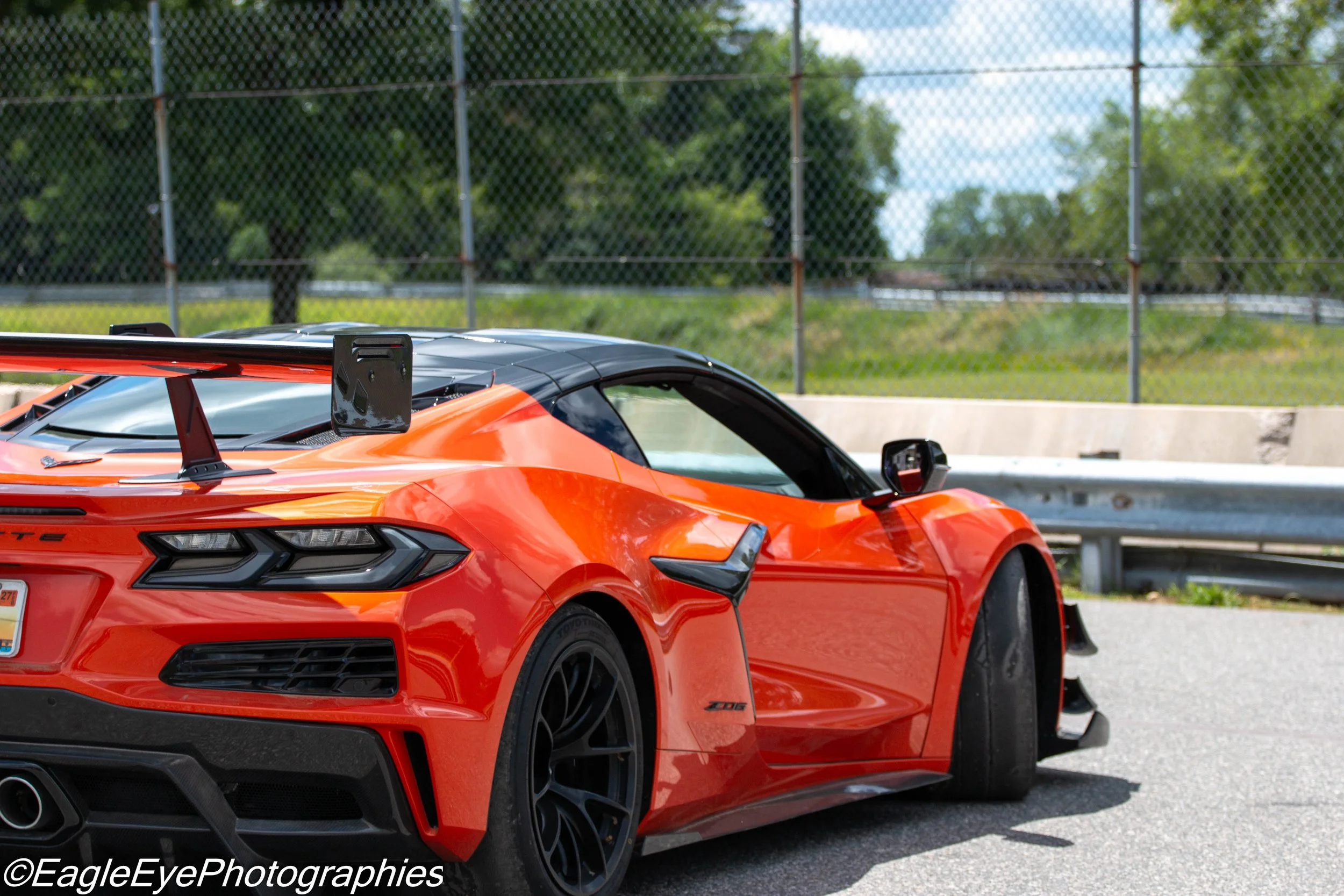 A bright orange sports car with a black spoiler parked near a chain-link fence with green trees in the background.