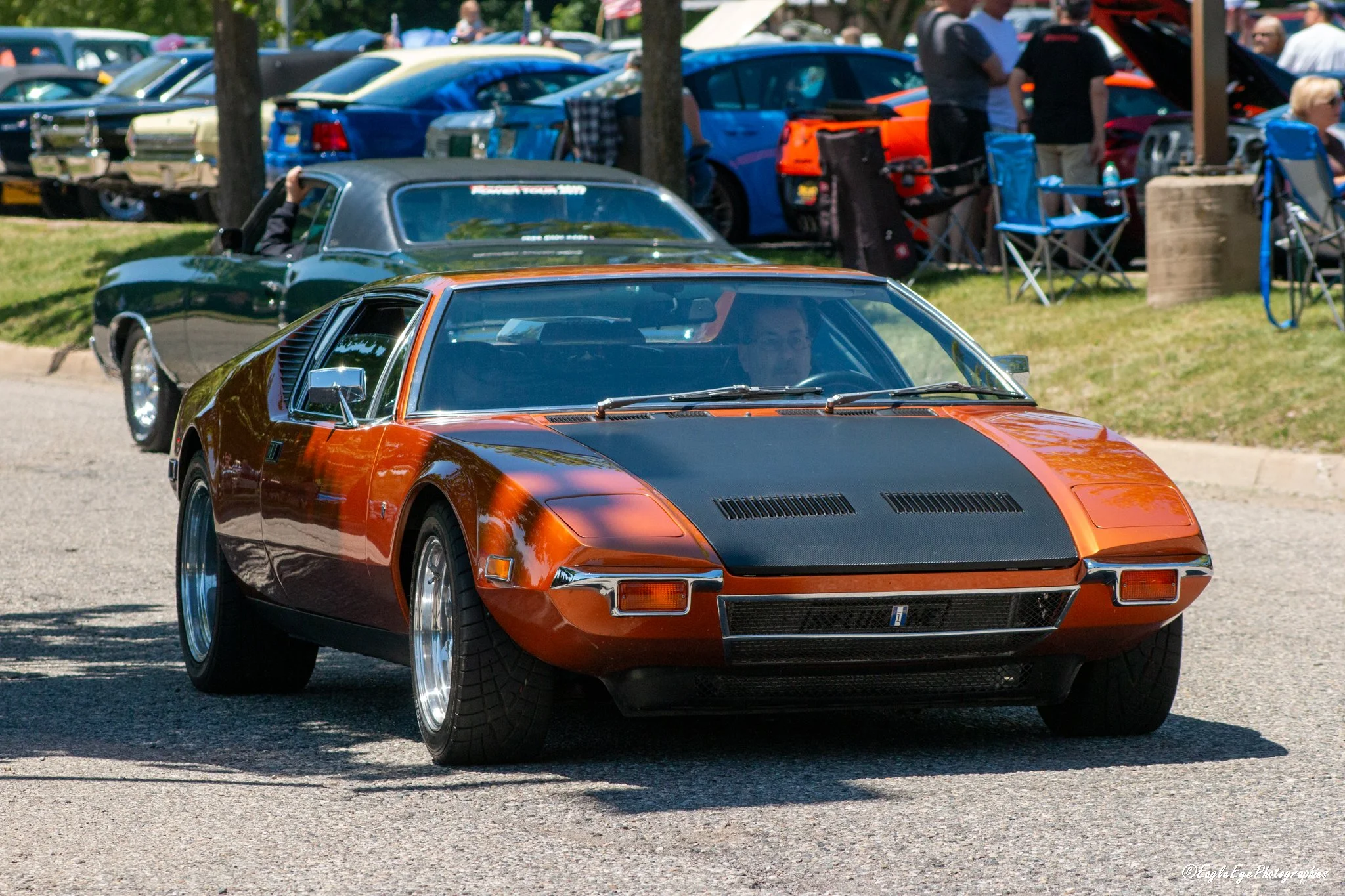 A classic orange and black sports car with pop-up headlights parked on a street at a car show, with other vintage cars and people in the background.