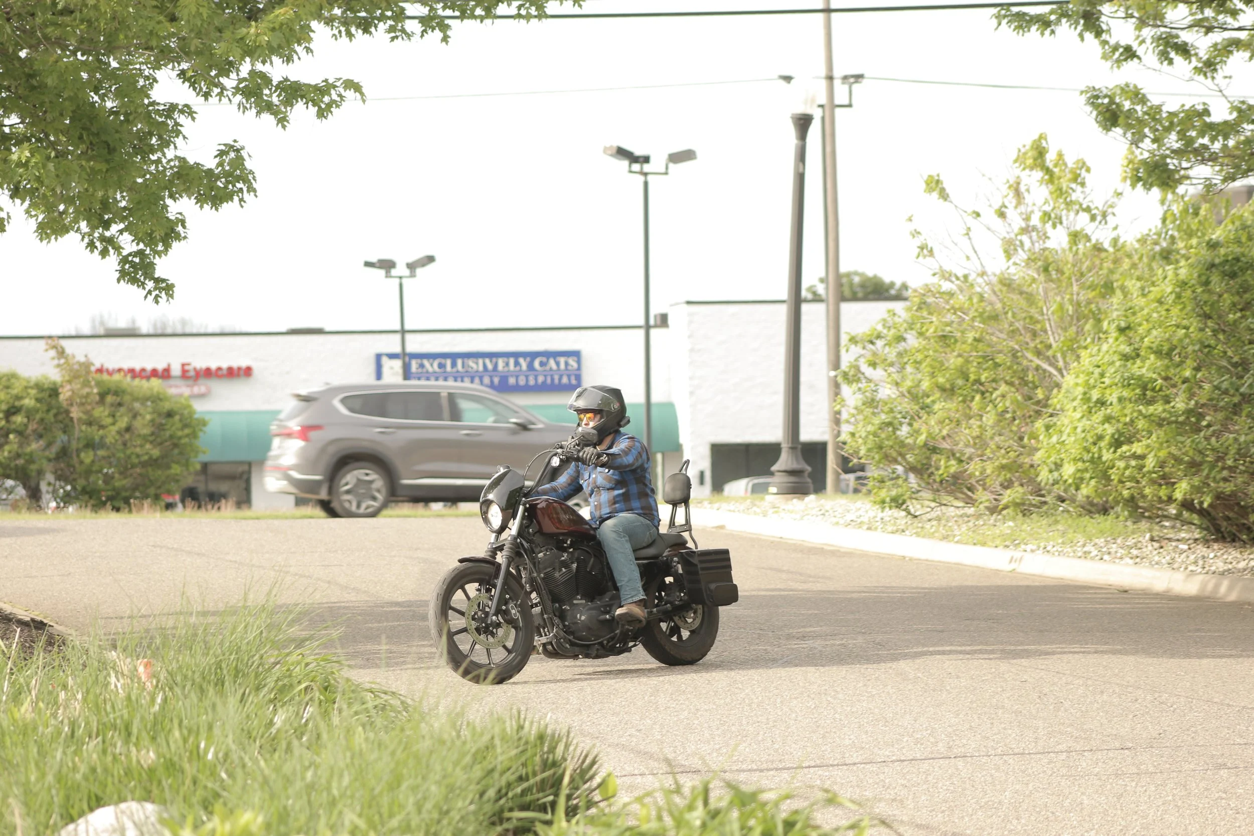 A man riding a motorcycle on a street, wearing a helmet and sunglasses, with trees and parking lot in the background.