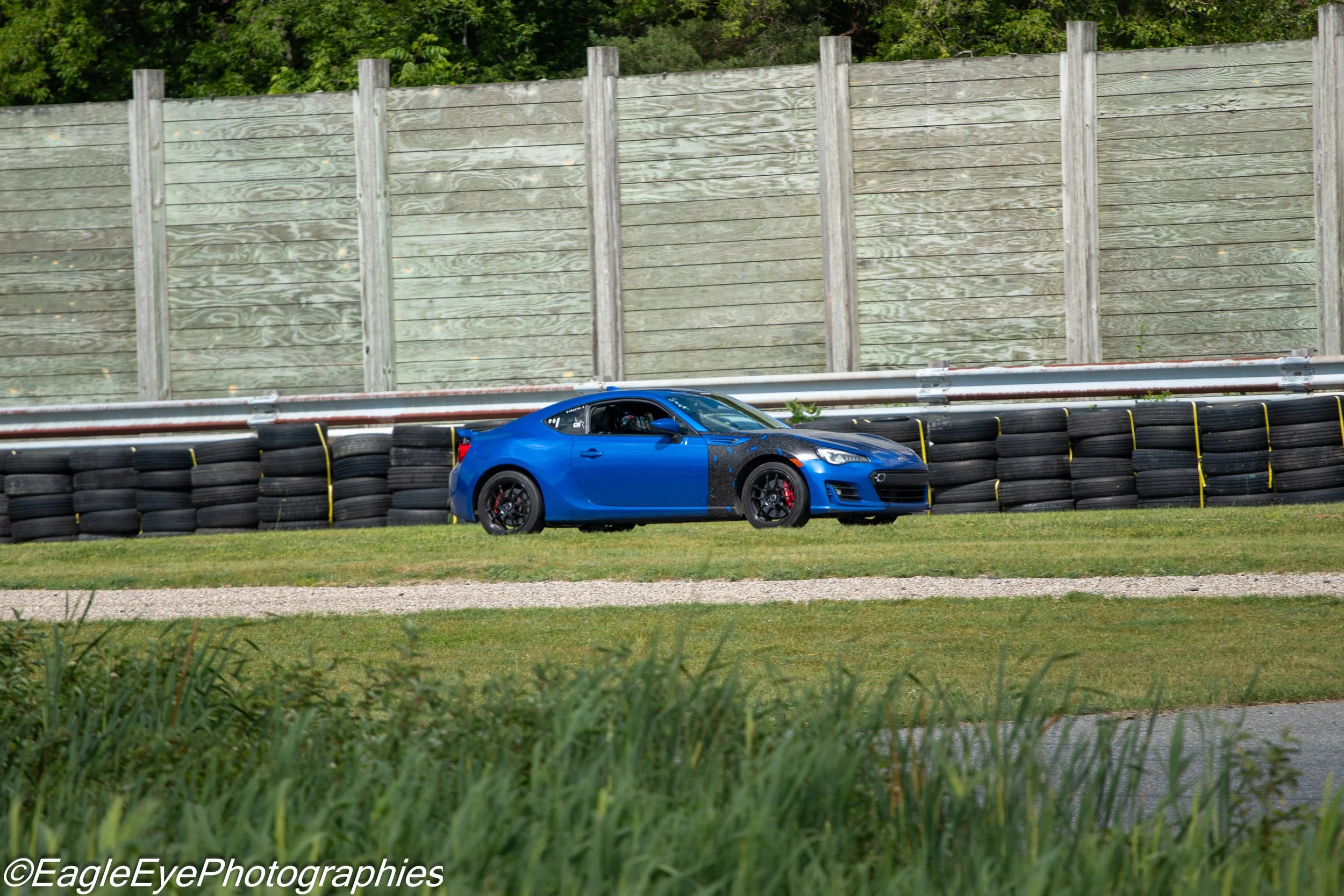 A blue sports car driving on a race track with a wooden fence and green trees in the background.