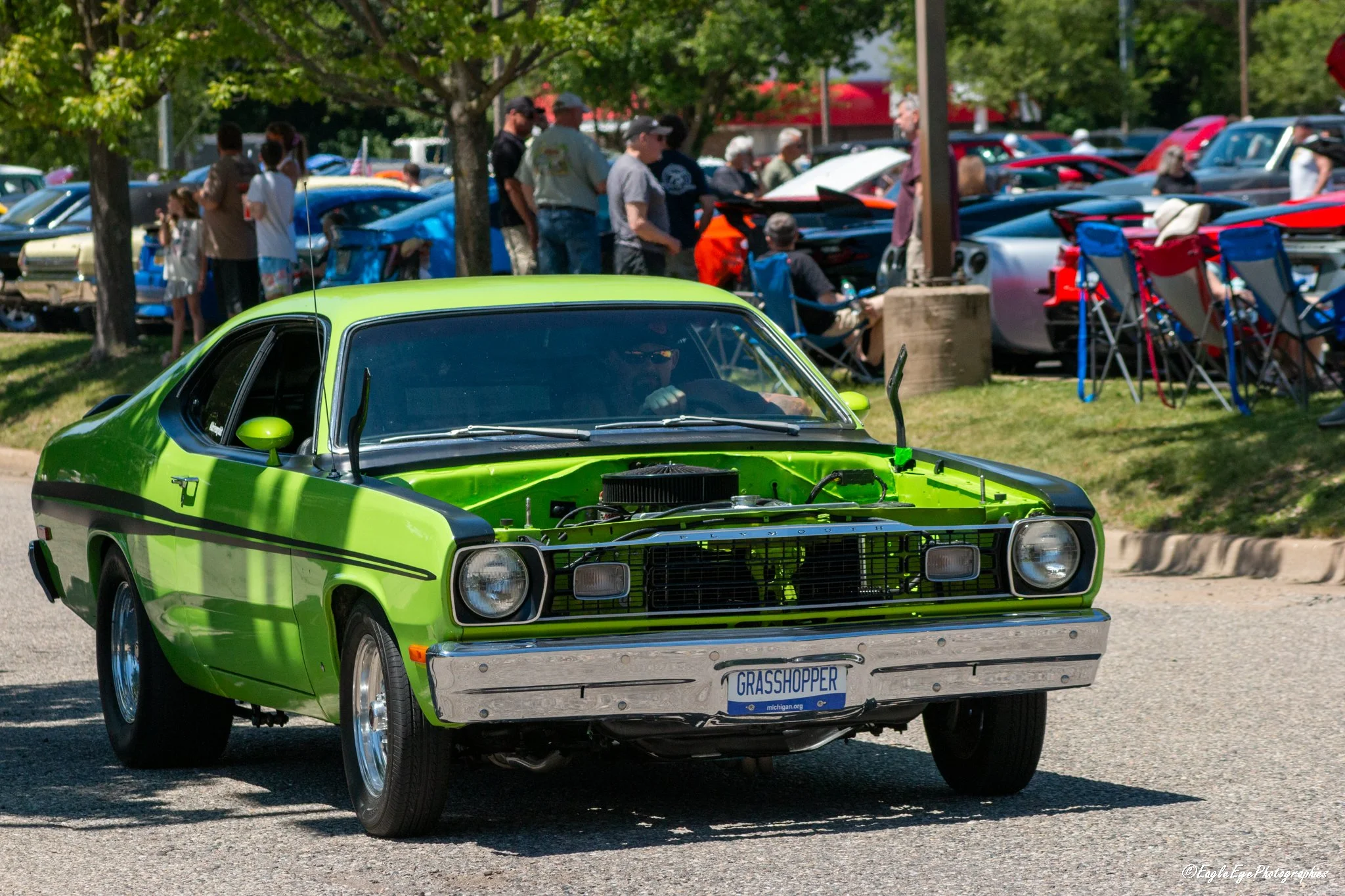 A bright green vintage car with a black grille and a Michigan license plate that reads 'GRASSHOPER' driving past a crowd at a car show, with many other cars and people in the background under trees.