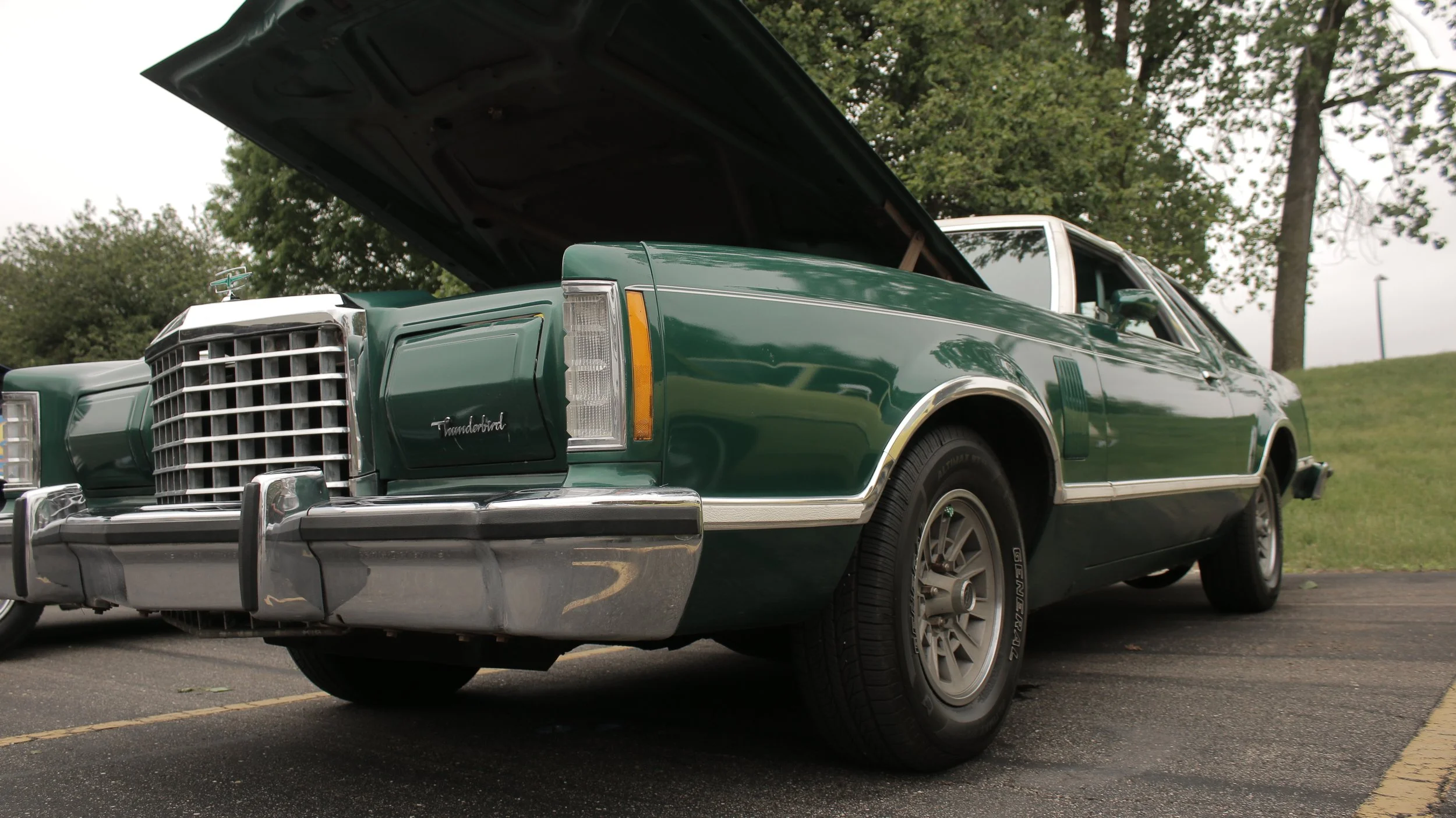 A vintage green Mercury Thunderbird car with its hood open, parked outdoors on a paved area with grass and trees in the background.