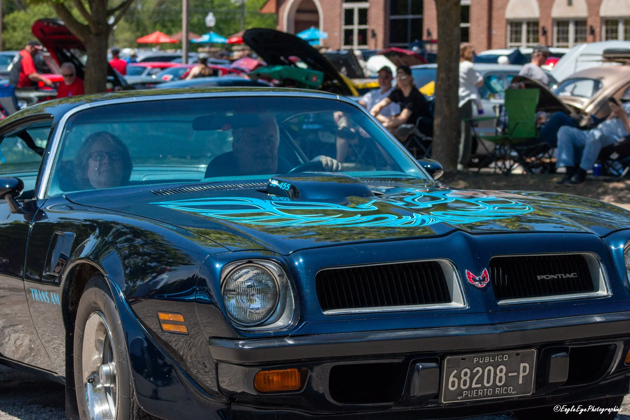 A black classic Pontiac Trans Am with blue flames on the hood, parked at a car show on a sunny day. In the background, people are sitting in lawn chairs, and other cars are on display with their hoods open.