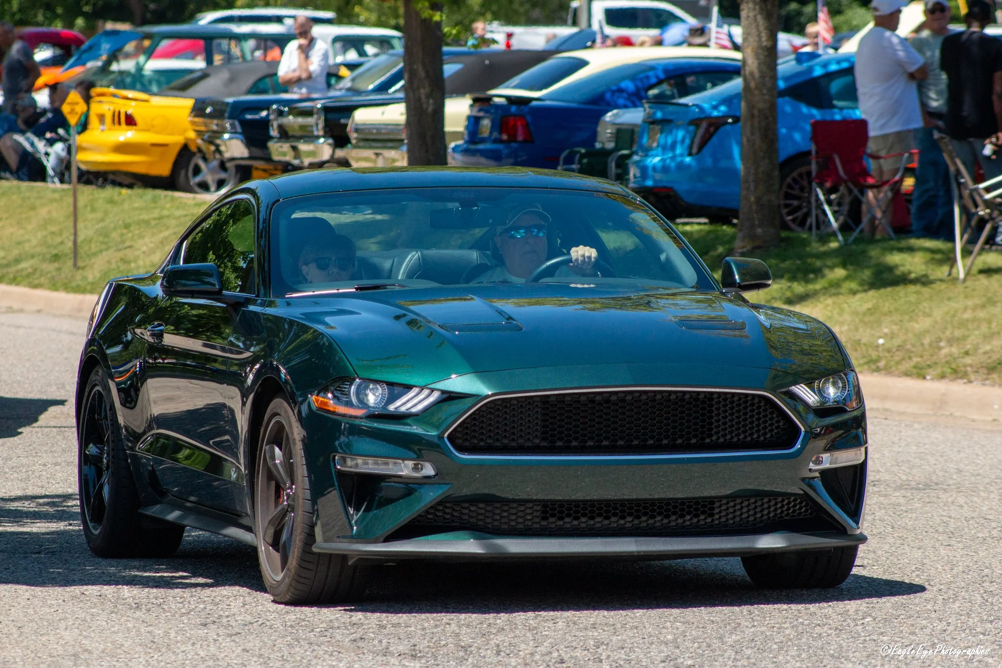 A black sports car on a paved road with people and parked cars in the background, some of whom are looking at the car. The scene appears to be at an outdoor car show or gathering.