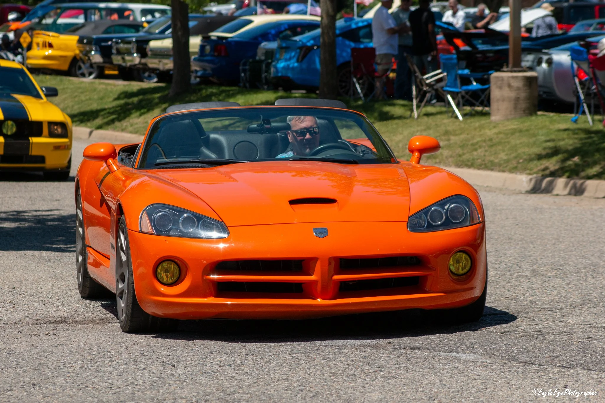 A bright orange sports car driving on a street with a large crowd of cars and people in the background.