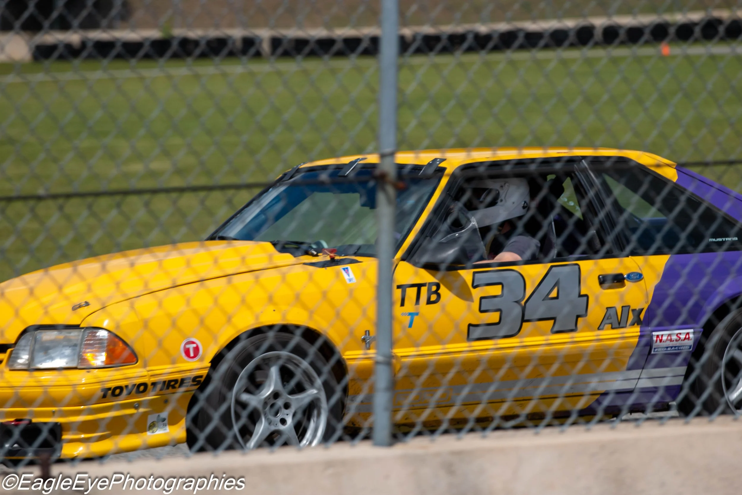 A yellow race car behind a chain-link fence at a racetrack.