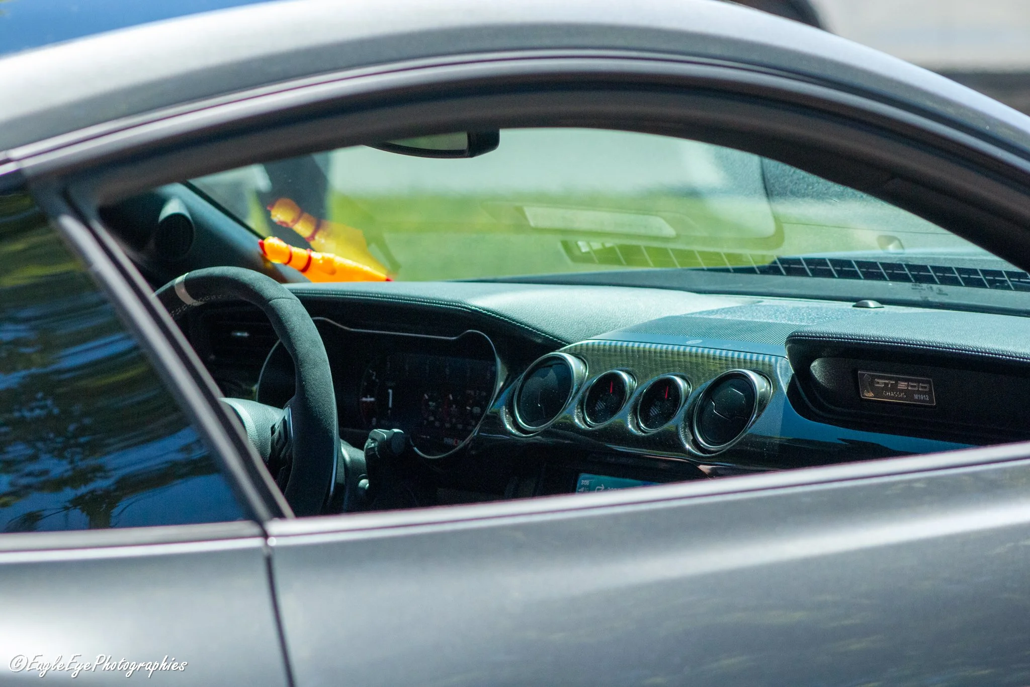 The interior of a car showing a black steering wheel, a dashboard with circular air vents, and a dashboard plaque reading 'GT350 R'. The windshield has a yellow mannequin arm hanging from the rearview mirror, and the image is taken through the car's 