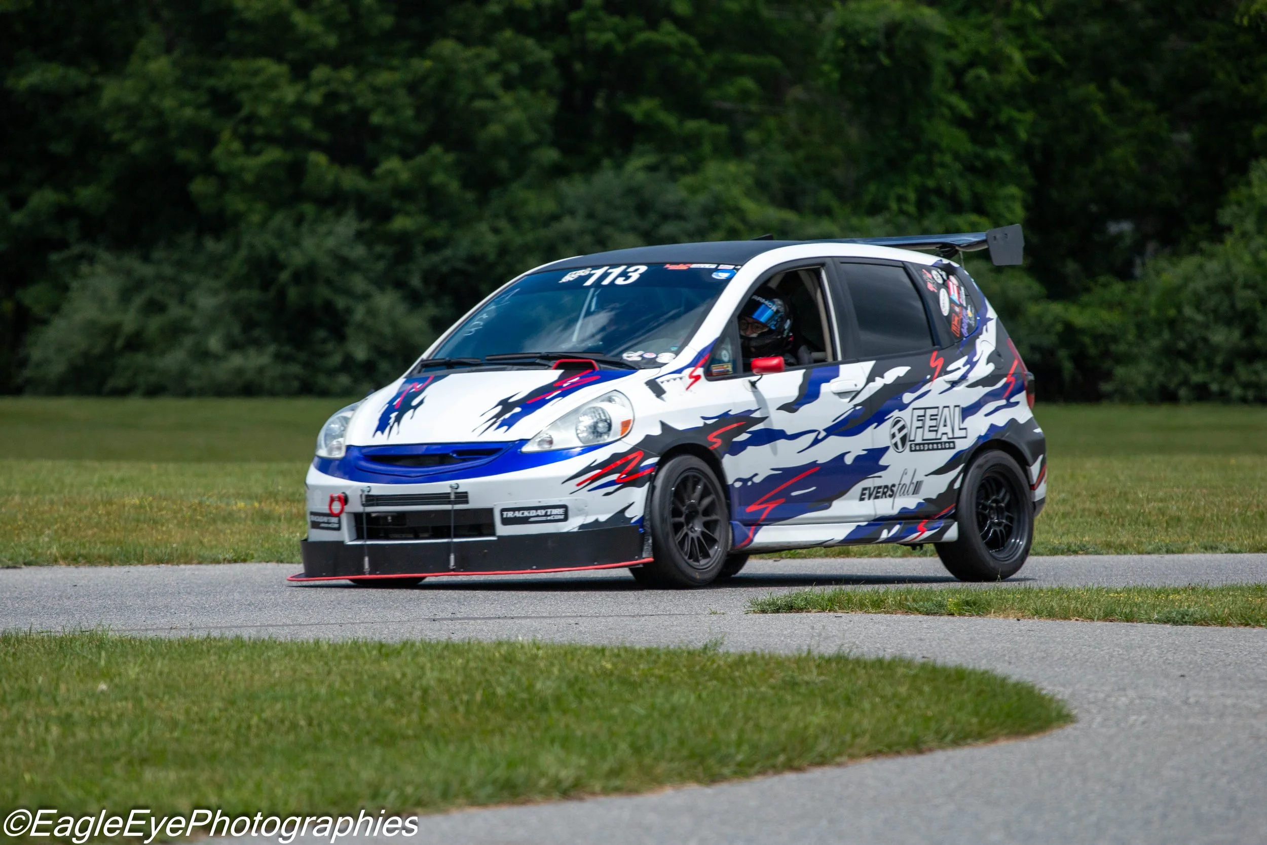 A race car with a dragon graphic and sponsorship stickers, driven by a person wearing a helmet, on a grassy and paved area with a background of green trees.