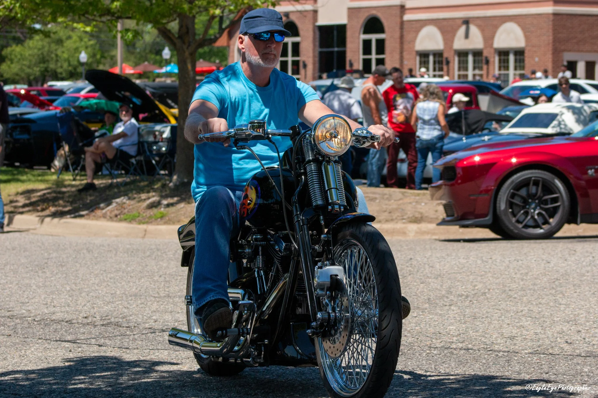 A man with a gray beard wearing sunglasses and a blue cap riding a motorcycle at a car show or gathering against a background of parked cars and people.