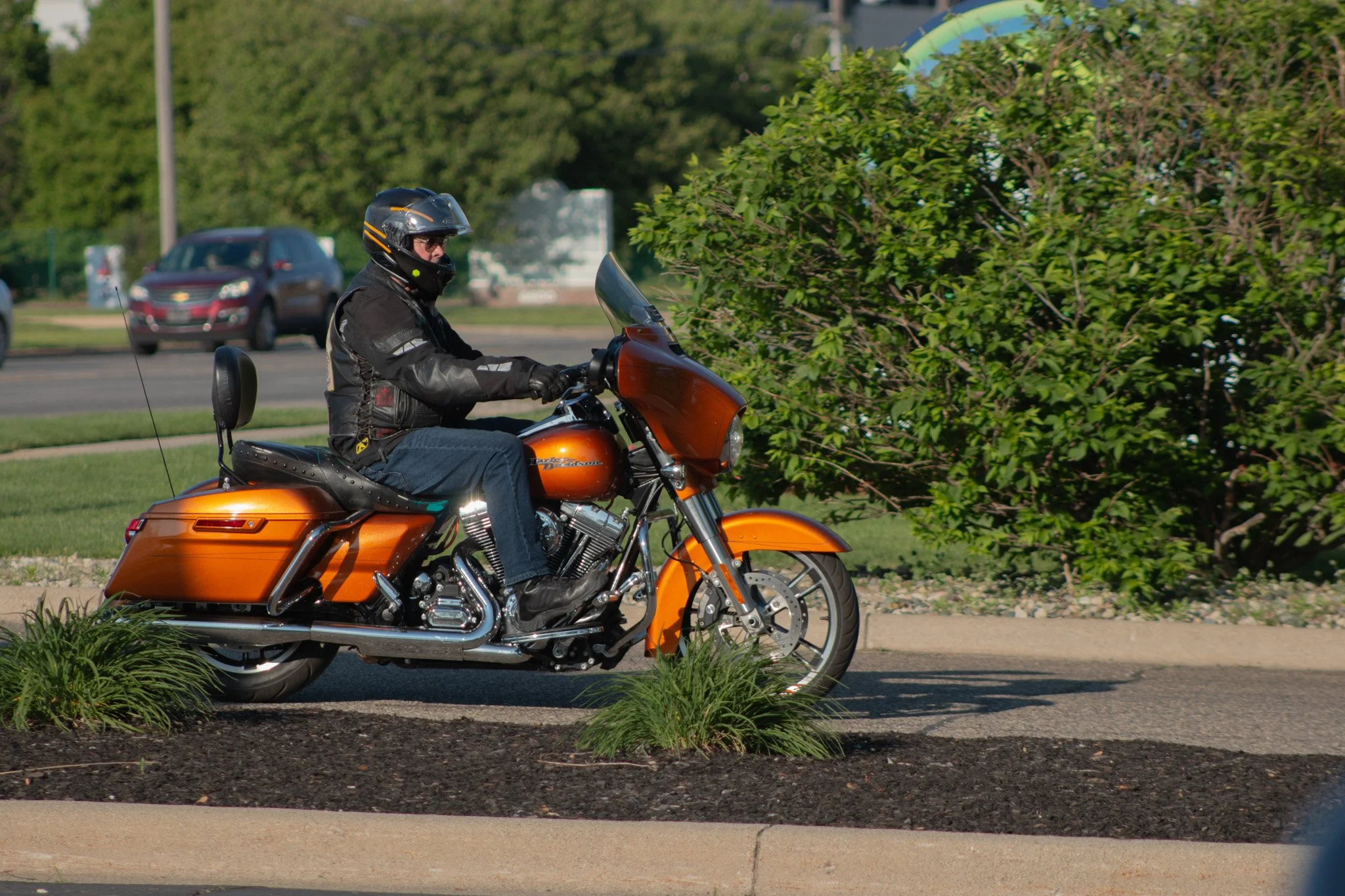 A person riding an orange motorcycle on a paved road near a grassy area with bushes, wearing a black helmet and black leather jacket.