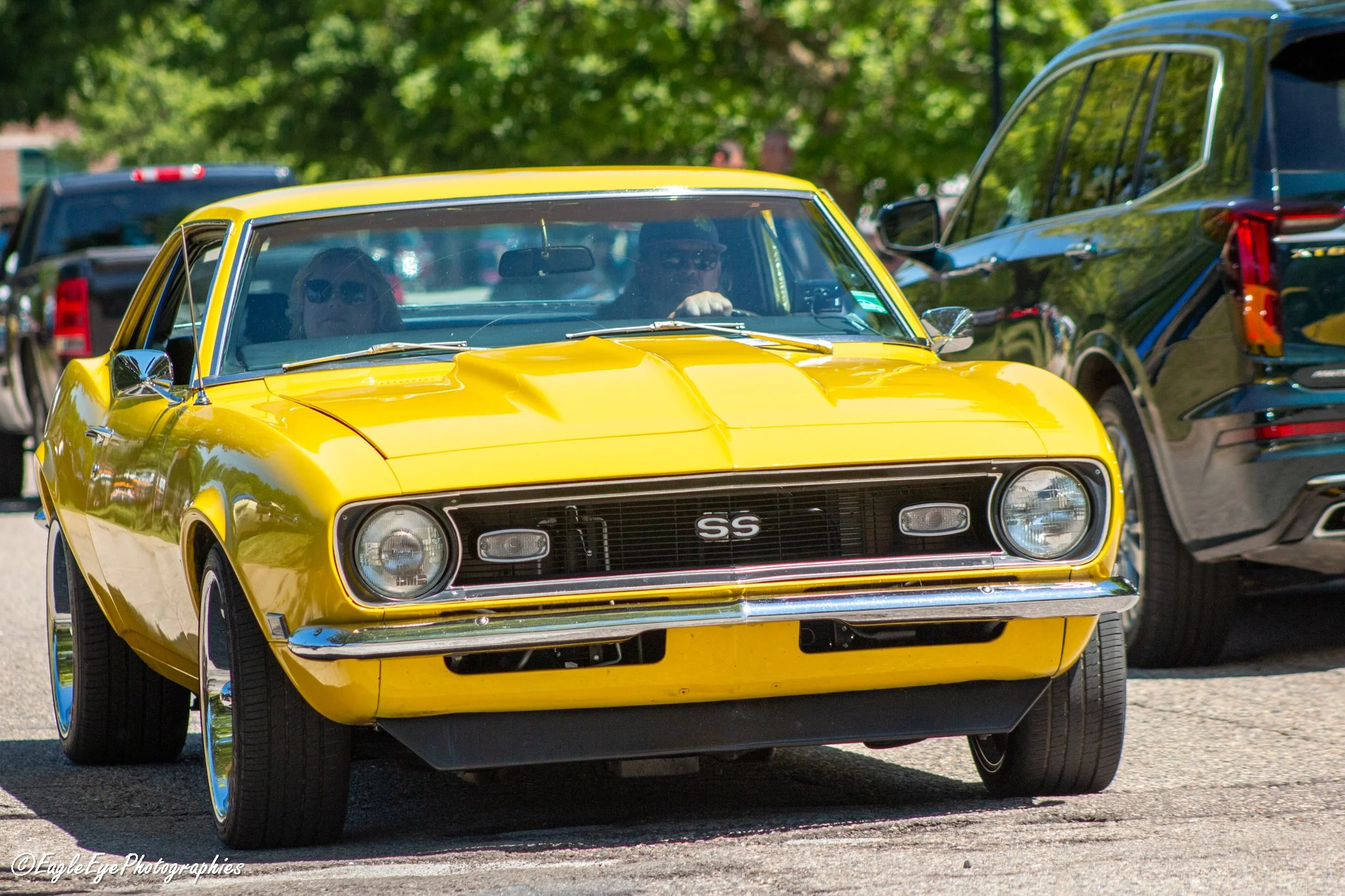 A bright yellow vintage Chevrolet Camaro SS with black racing stripes parked outdoors, with people inside the car and parked vehicles and trees in the background.