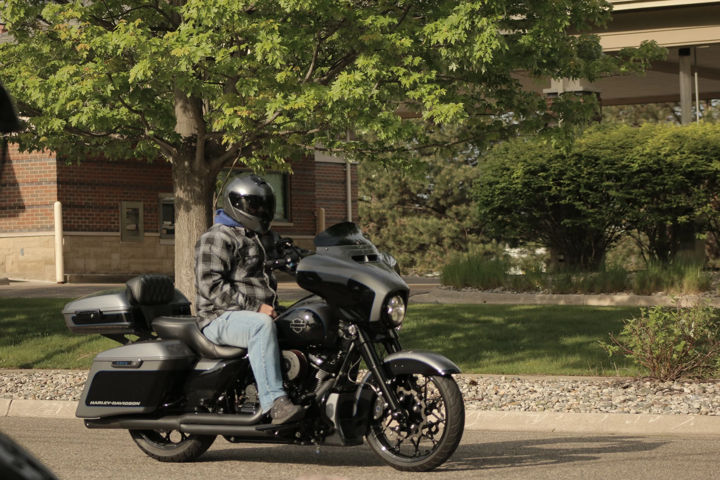 A person wearing a helmet and plaid shirt riding a black Harley-Davidson motorcycle on a street with green trees and a brick building in the background.