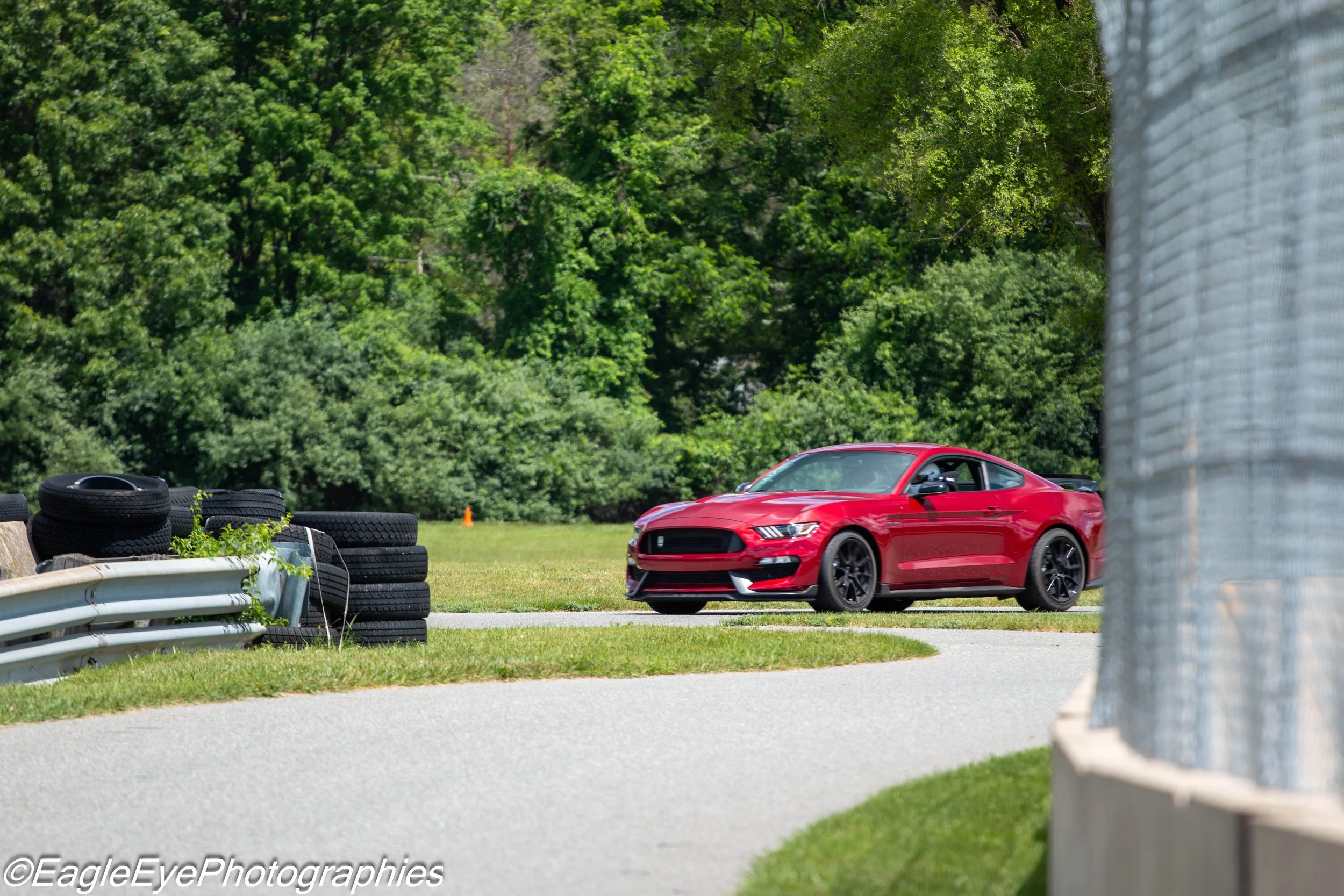 A red sports car driving on a race track with tire stacks and a safety barrier in the foreground, surrounded by green trees.