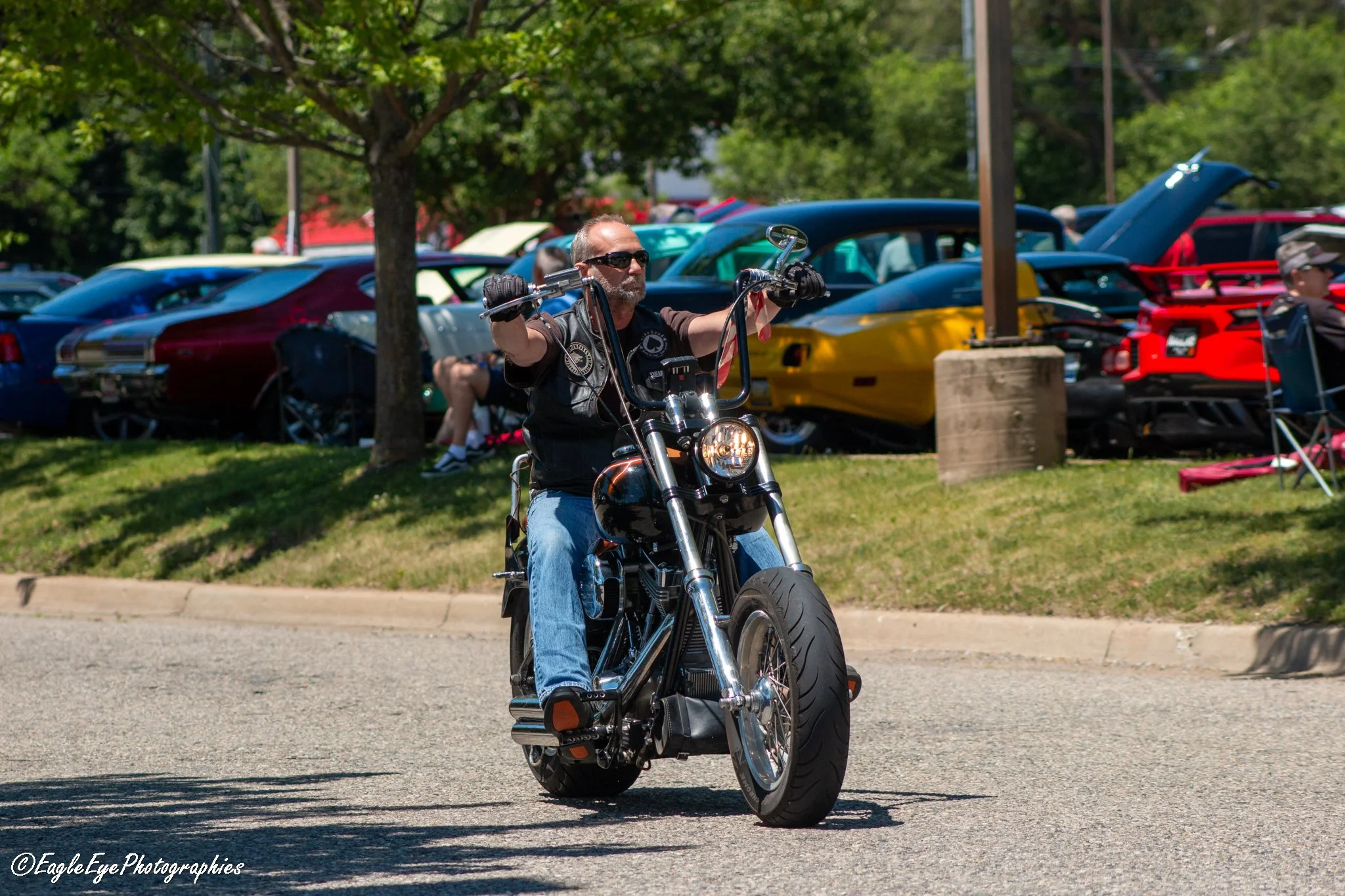 A man riding a black motorcycle on a sunny street with cars parked on the side and green trees in the background.