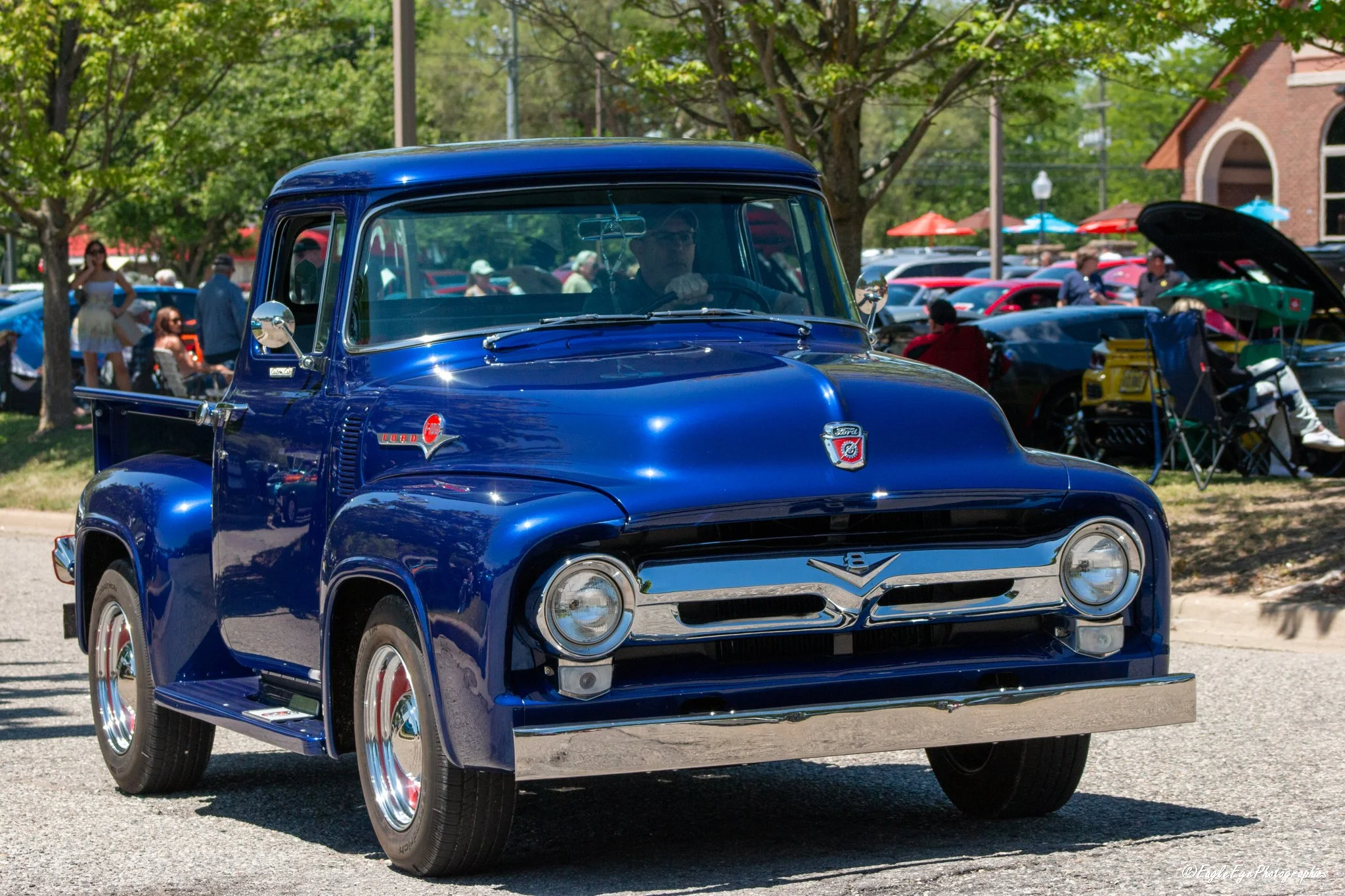 A vintage blue Ford pickup truck at a car show with people in the background and trees providing shade.