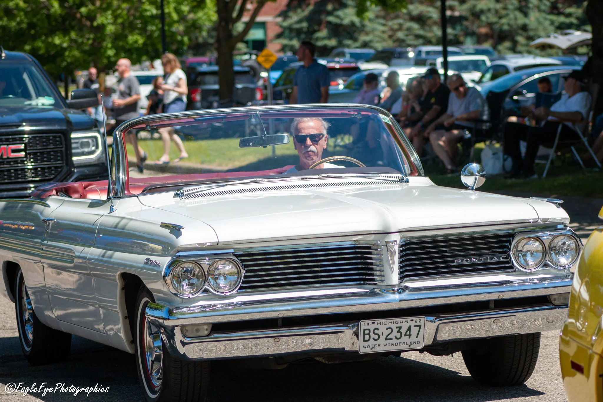 A vintage white convertible Pontiac car with Michigan license plate BS-2347, parked at an outdoor car show. In the background, a man with sunglasses and gray hair is sitting inside the car. Several people and modern vehicles are visible, along with t