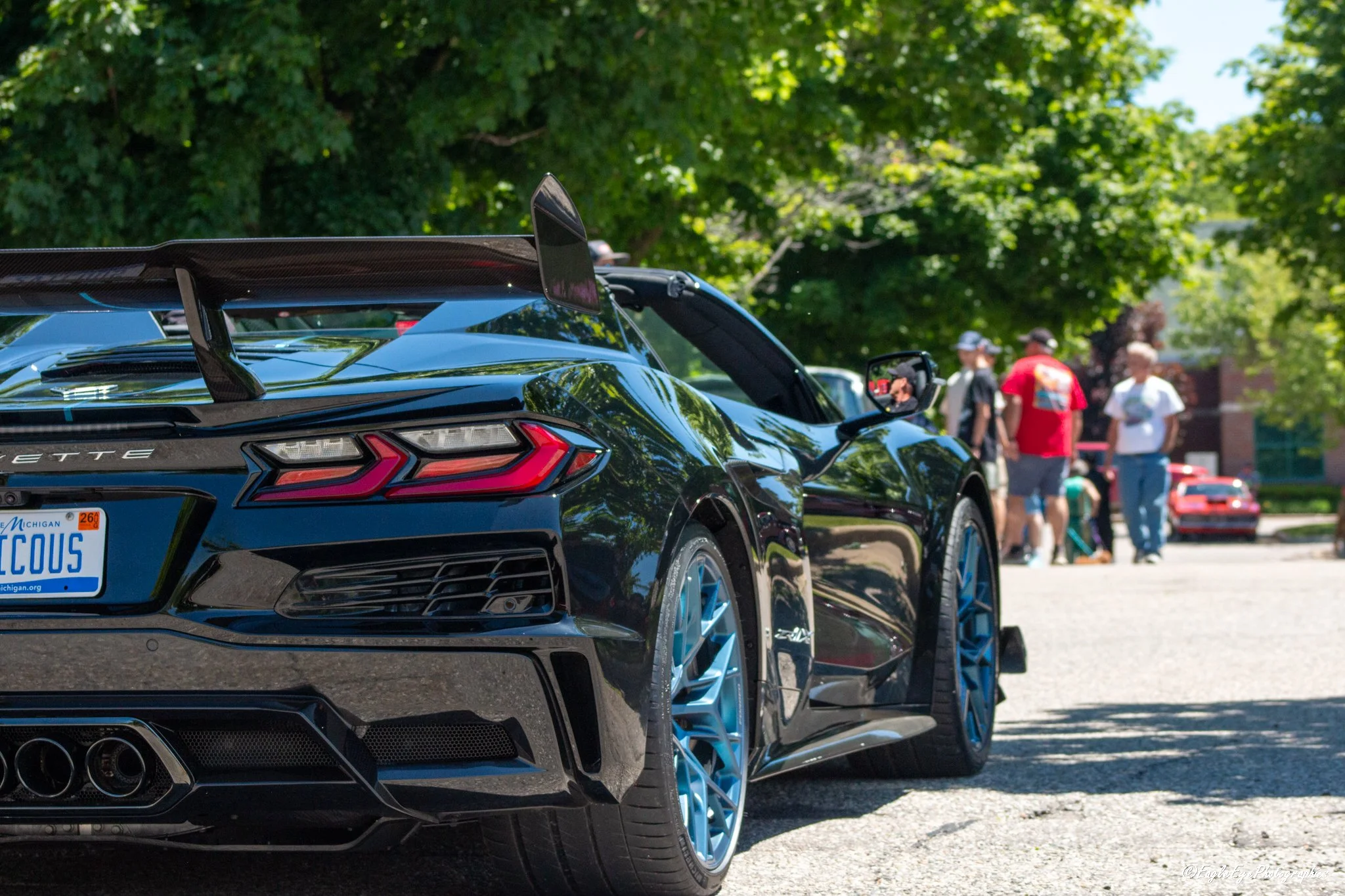 Close-up of a black sports car with carbon fiber rear wing and blue accents, parked outdoors with trees and people in the background.
