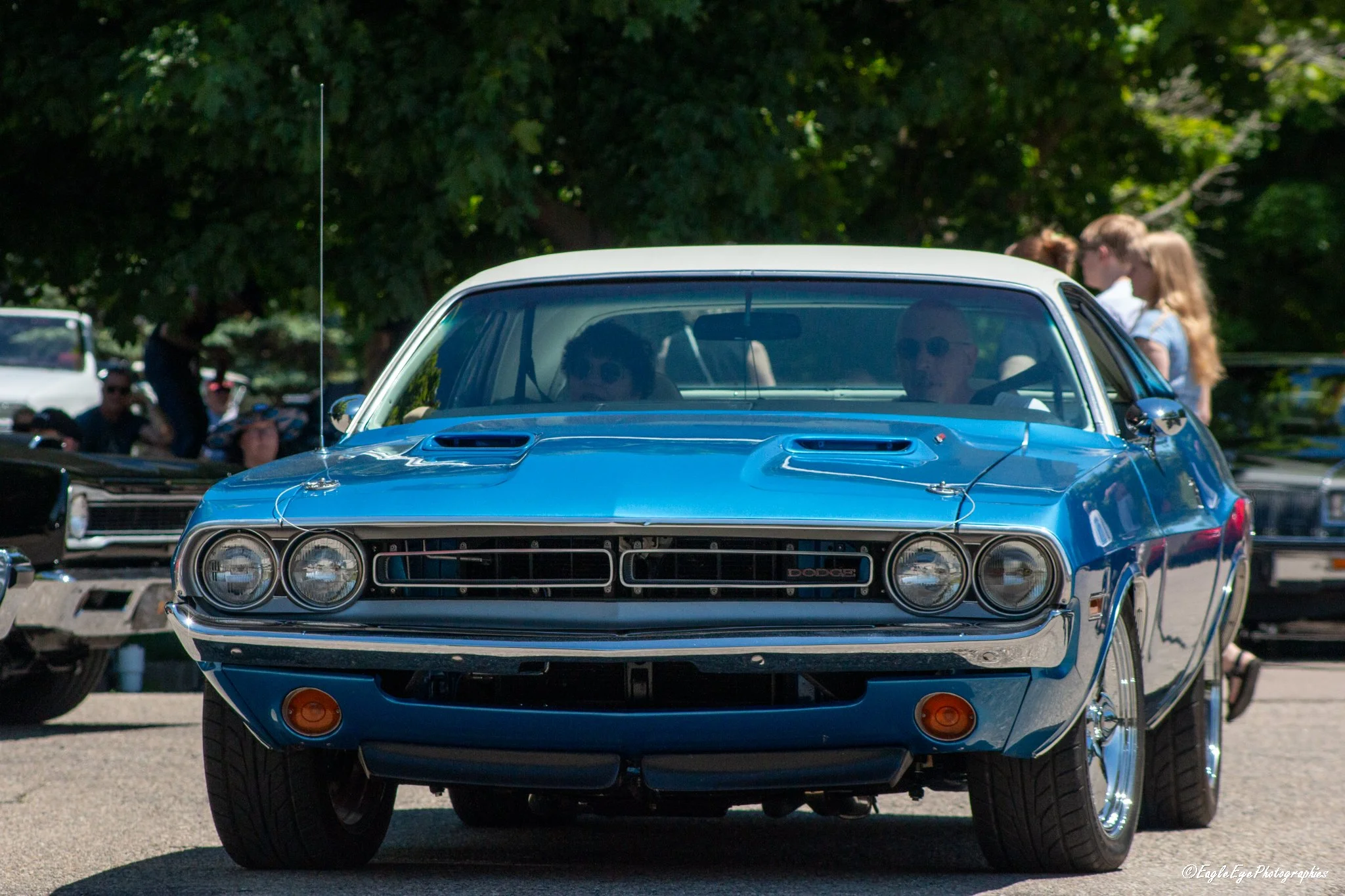 A blue vintage Dodge car parked outdoors with trees in the background and people, including women with blonde hair, in the distance.