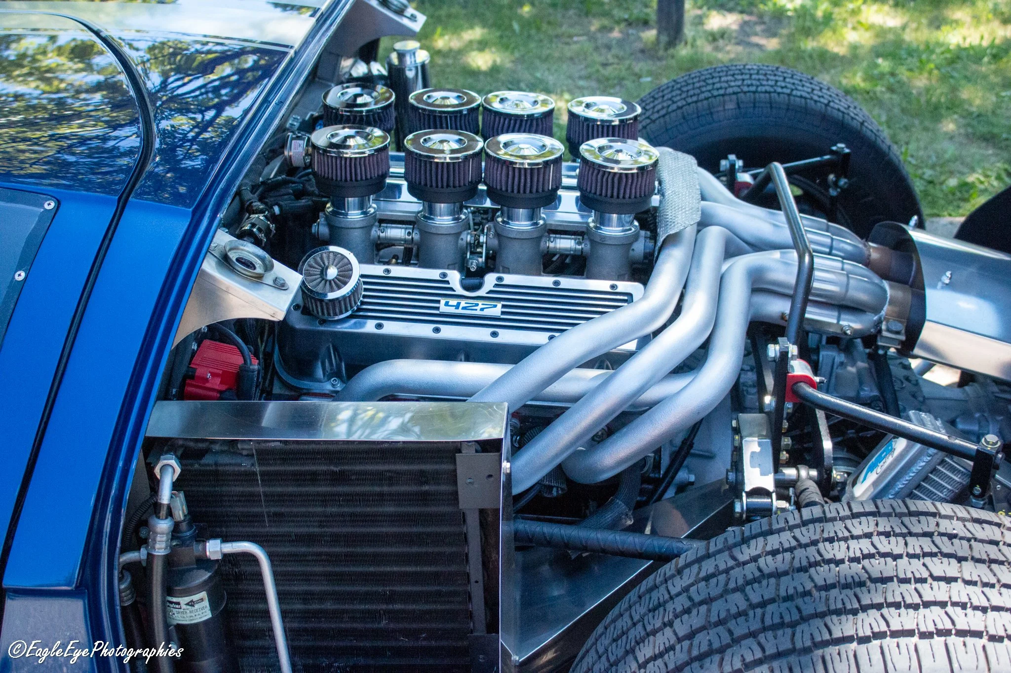 Close-up of a high-performance car engine with multiple air filters, visible exhaust pipes, and a tire in the background.
