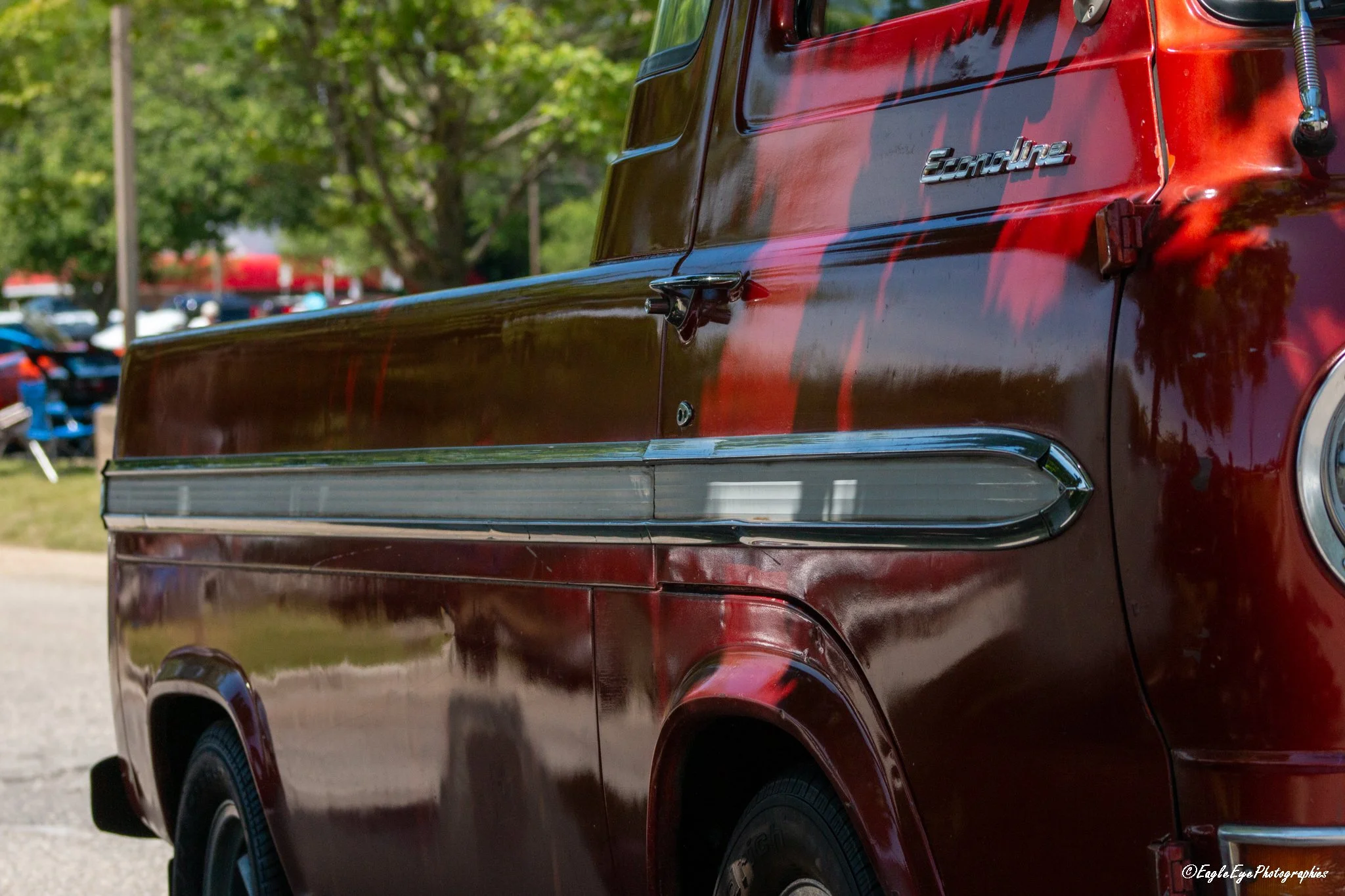 Close-up of a red vintage truck with a shiny exterior, showing the side with a chrome handle and a 'GMC' badge, parked outdoors with green trees and other vehicles in the background.