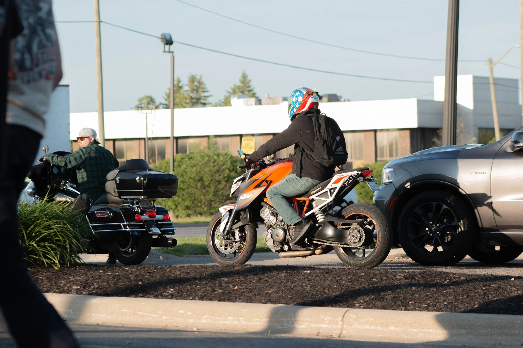 View of a motorcyclist with a colorful helmet riding a KTM motorcycle in traffic, with a man on a black motorcycle and a silver car nearby, amid a suburban setting with power lines and trees in the background.