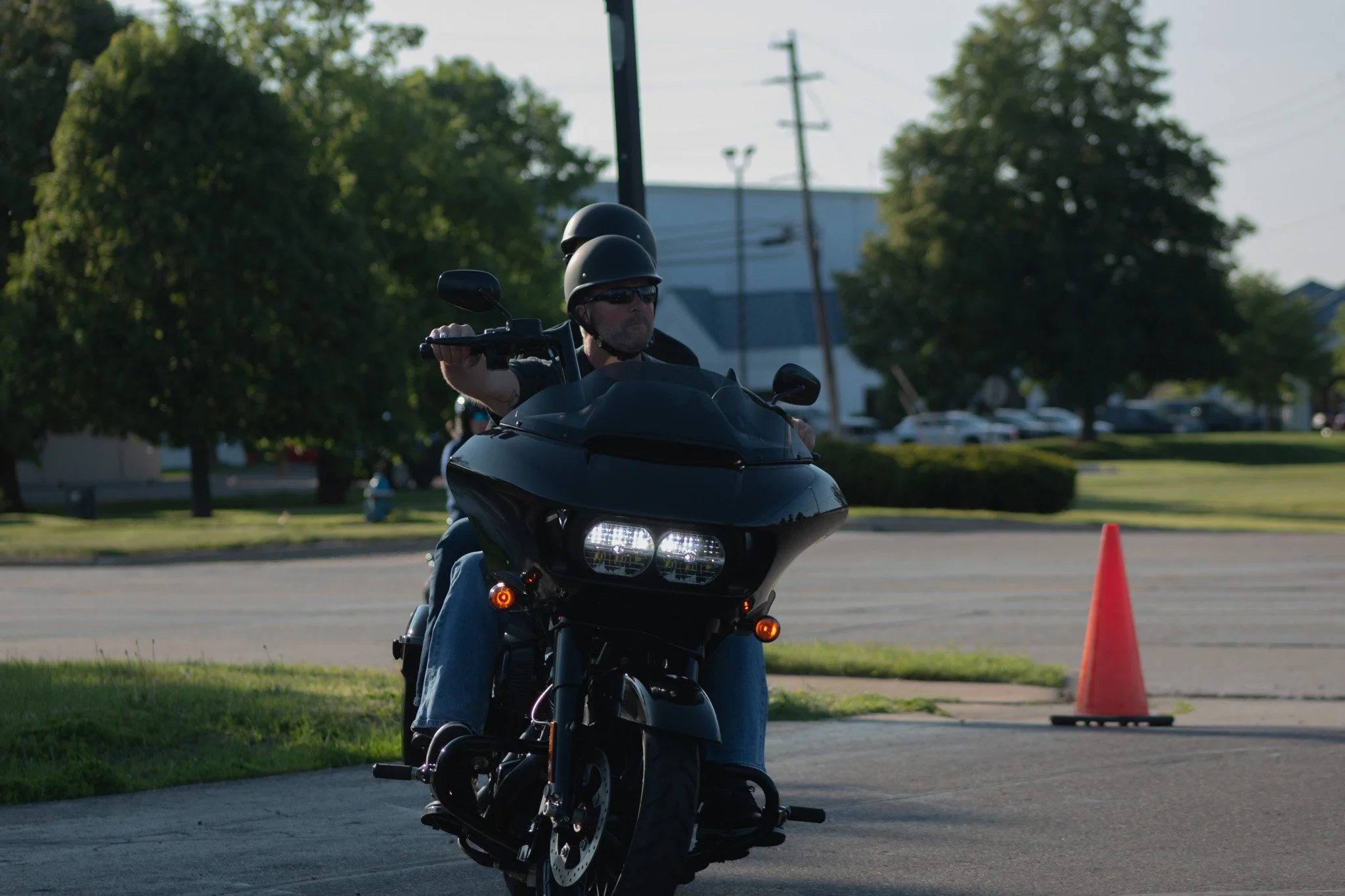 A man wearing a helmet and sunglasses riding a black motorcycle in an outdoor setting, with orange traffic cones nearby and trees in the background.