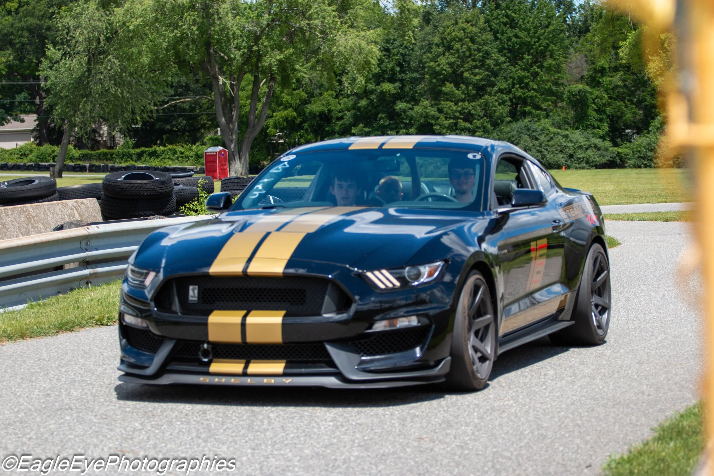 A black Ford Mustang Shelby with yellow racing stripes, driven by two men, on a curving race track with tires stacked along the side, surrounded by green trees and grass.