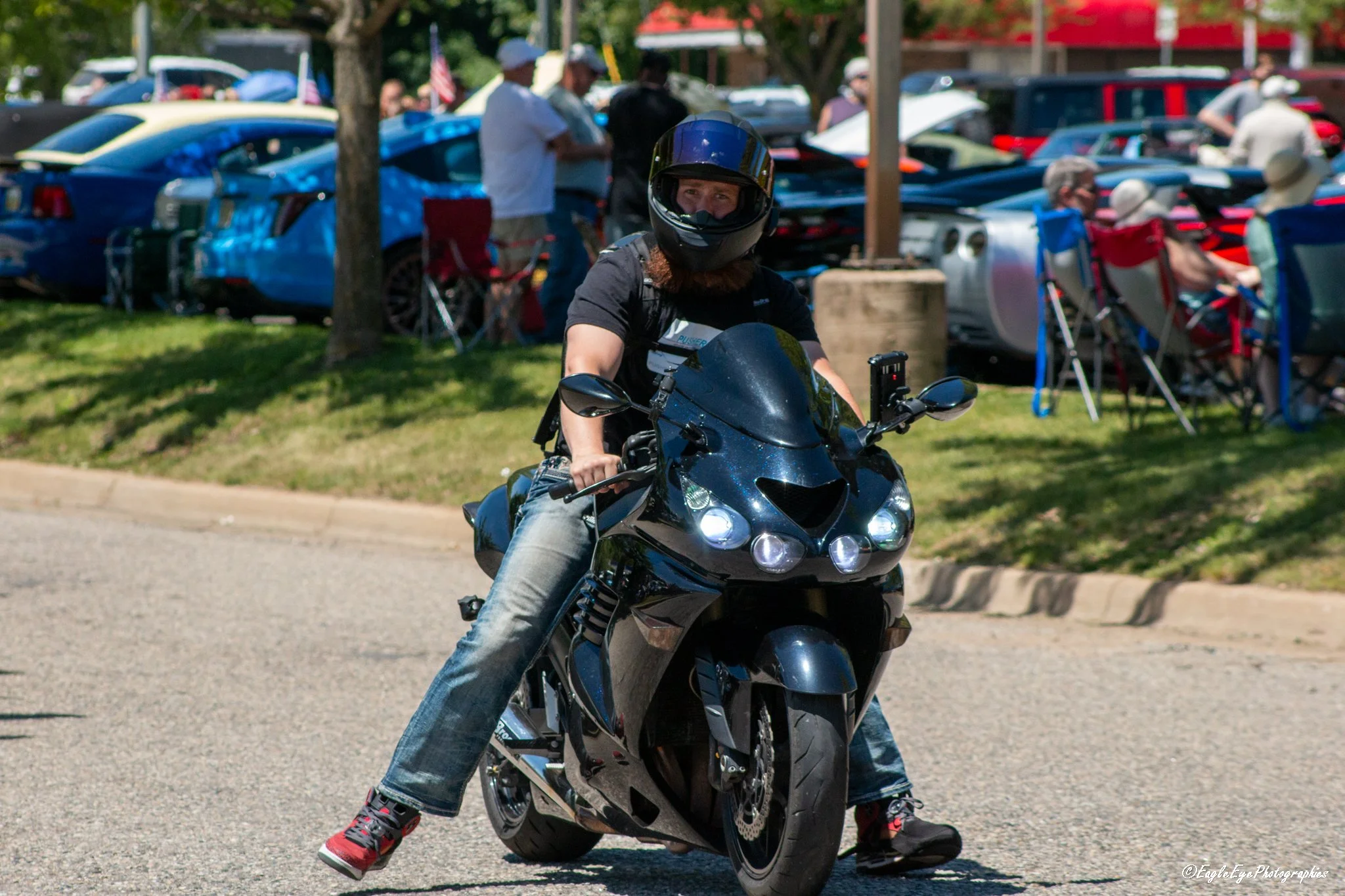 A person wearing a black motorcycle helmet riding a black motorcycle in front of a car show, with people looking at cars in the background.