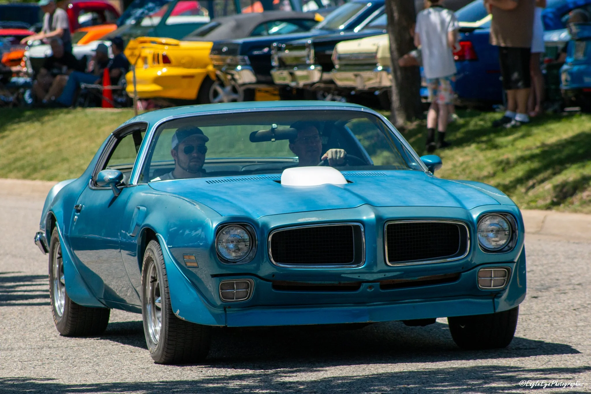 A blue classic muscle car with chrome wheels and a hood scoop, driving through a car show with other cars and people in the background.
