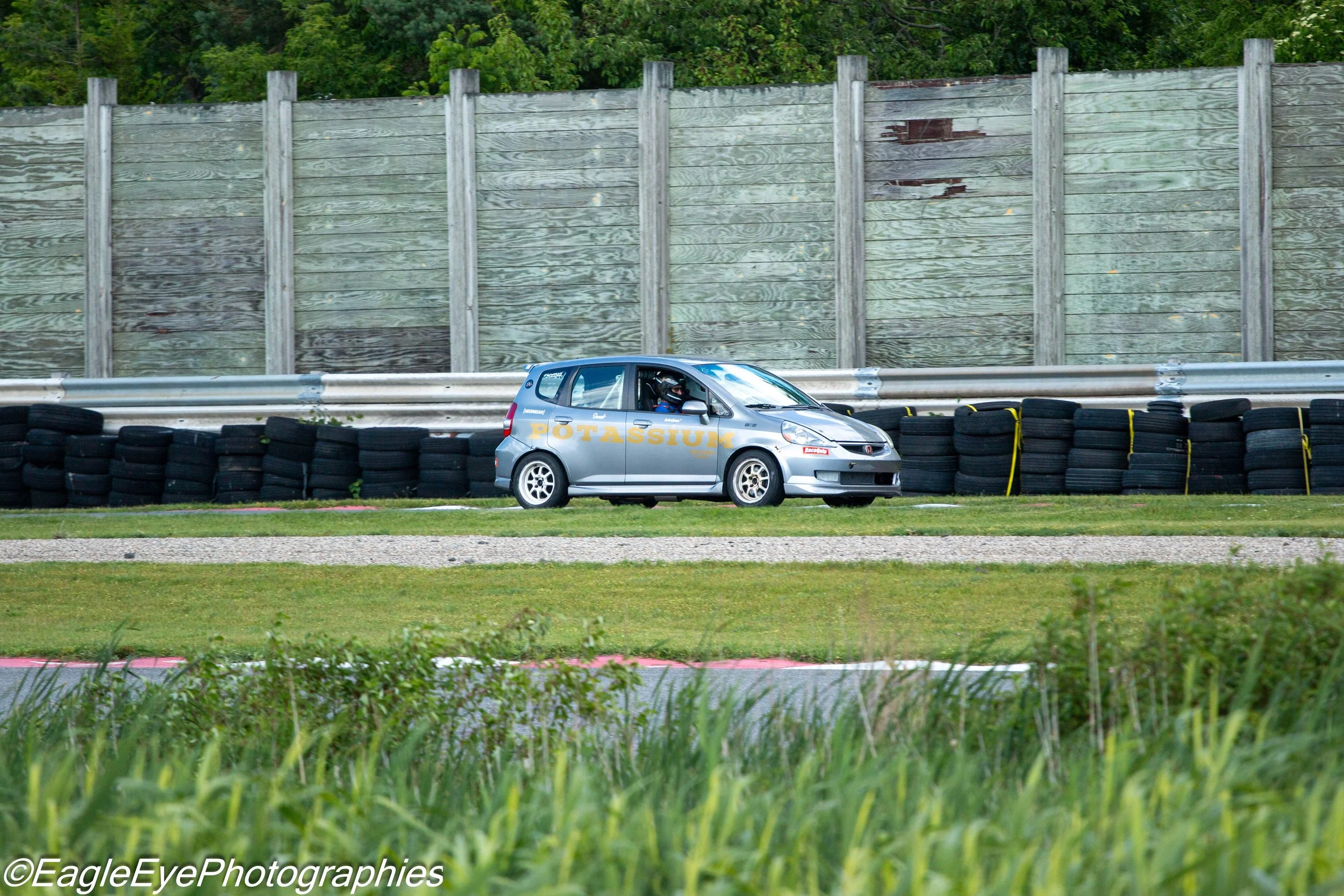 A silver rally car with the words 'POTASSIUM' on the side, driving on a race track behind a stack of black tires and a metal safety barrier, with a tall wooden fence and green trees in the background.