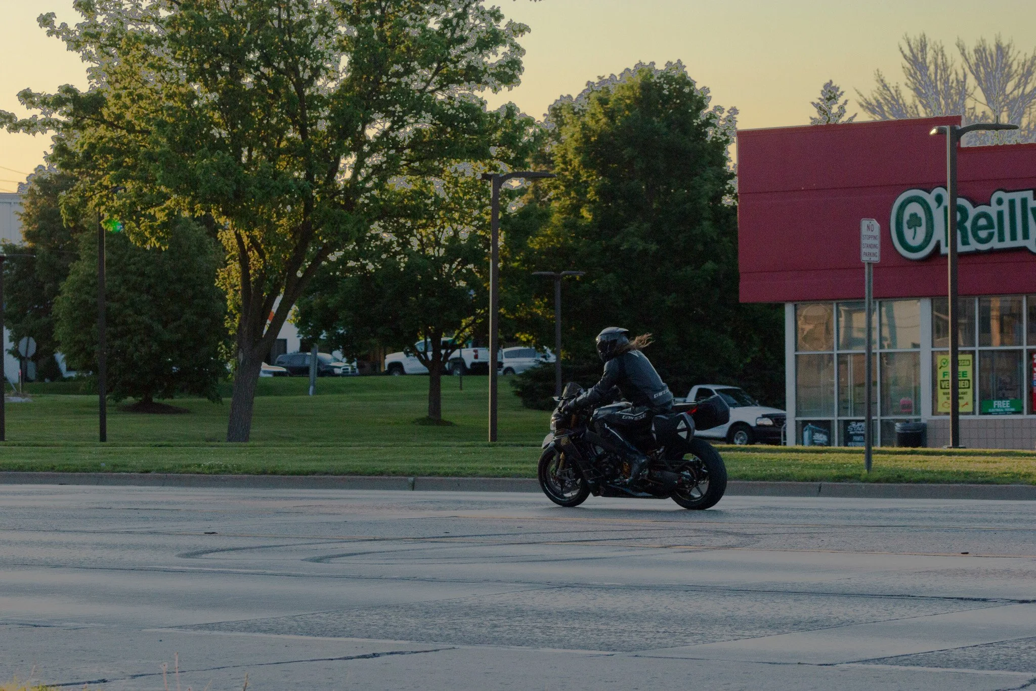 A motorcycle rider wearing a helmet and jacket riding a black motorcycle on a city street during sunset. Trees and parked cars are visible in the background, along with a red building with a sign reading "O'Reilly." The scene is illuminated by the wa