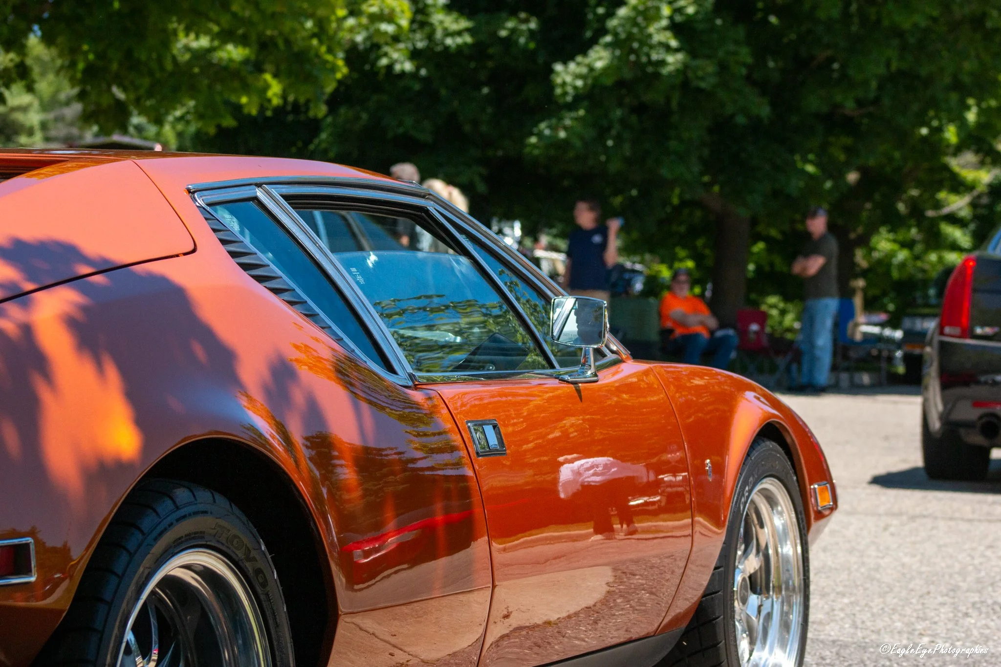 Close-up of a vintage orange sports car with chrome details, parked outdoors with trees in the background and people standing and sitting nearby.