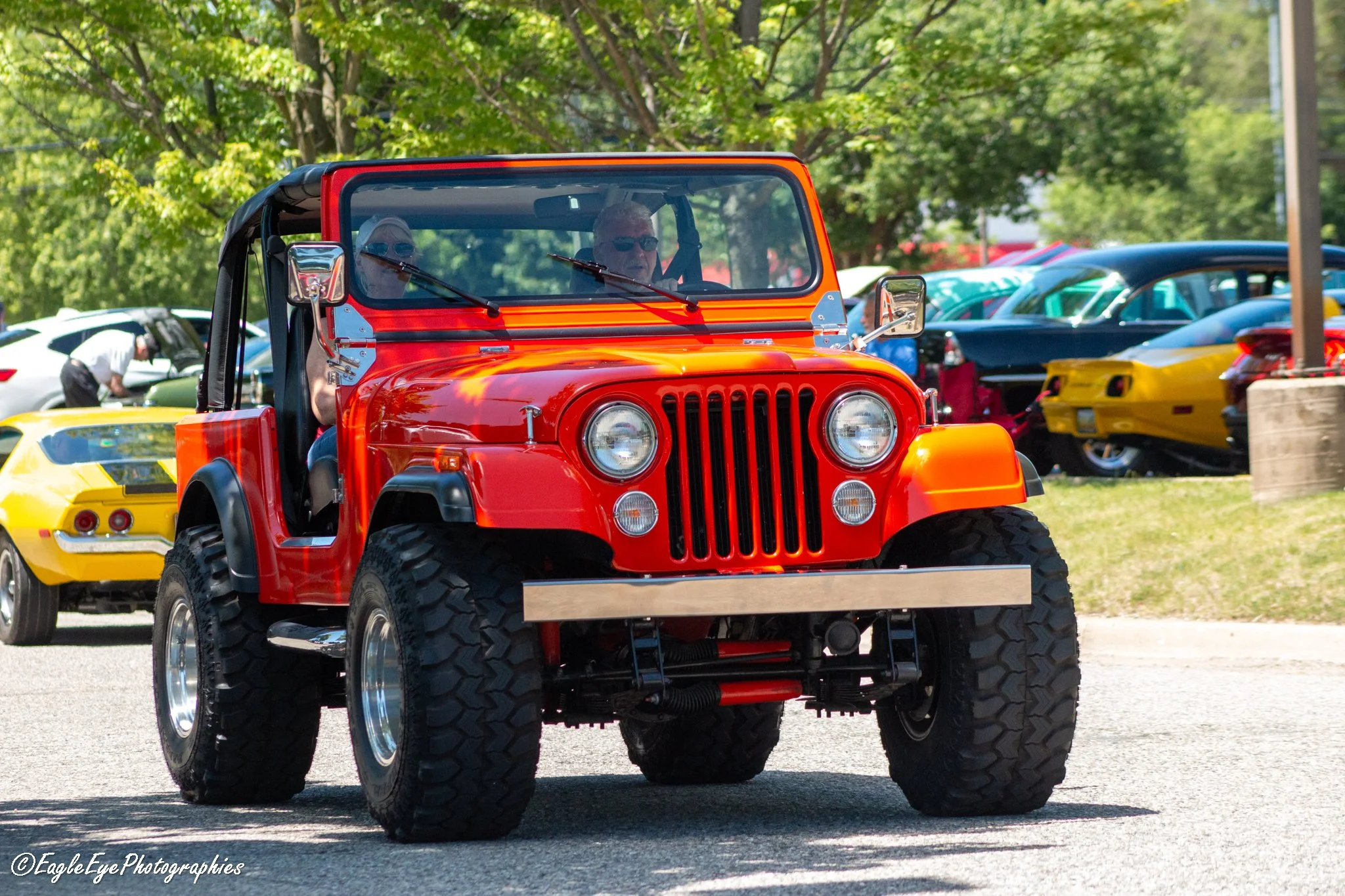 A bright orange vintage Jeep or off-road vehicle with large tires at an outdoor car show, with people and colorful cars in the background.
