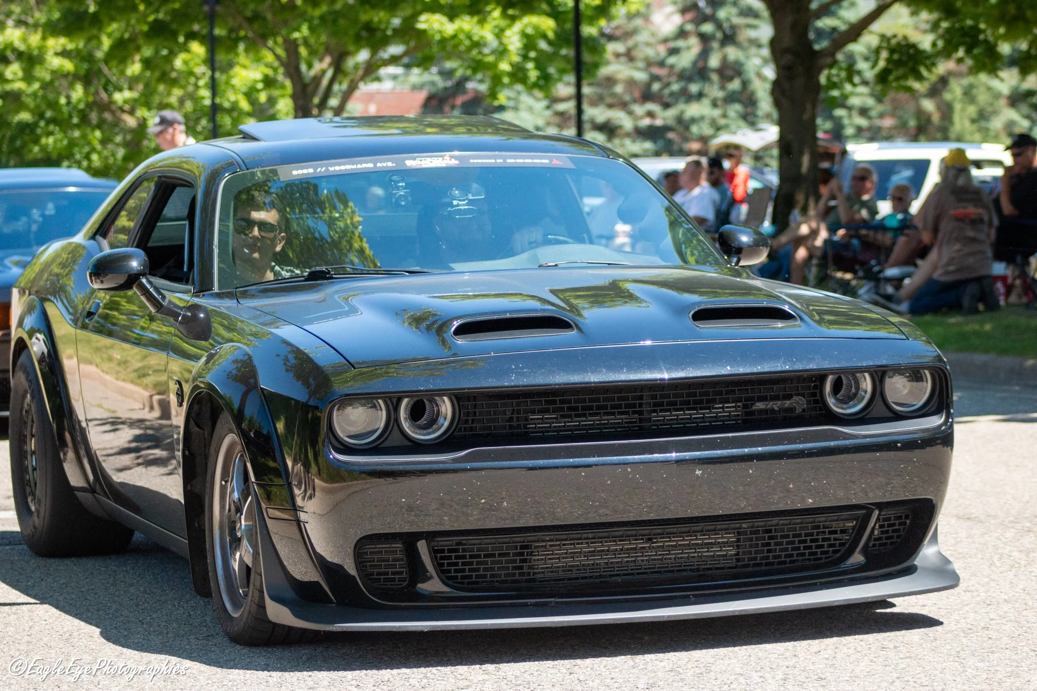 Close-up of a black sports car with chrome accents, parked outdoors with a reflection of trees on its surface. In the background, people are gathered, some sitting and some standing, in a park setting.